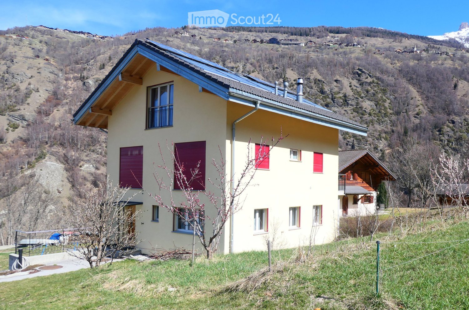 Two-story house with yellow walls, red shutters, and a blue roof, situated in a mountainous area with a view of the surrounding landscape.