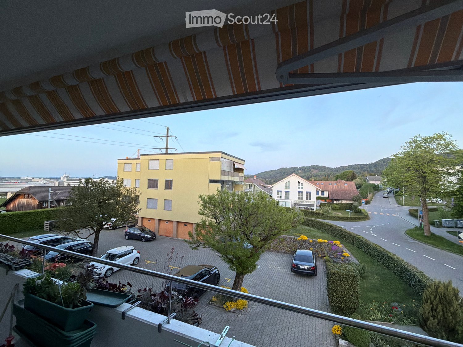 balcony with plants, view of a parking lot with cars, yellow building, trees, road