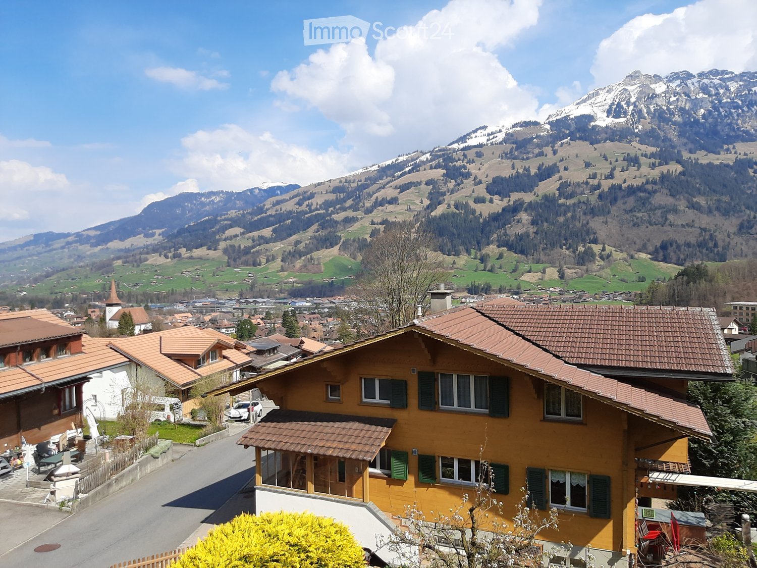 Swiss-style house, brown roof, yellow walls, multiple windows, surrounded by greenery, mountain view