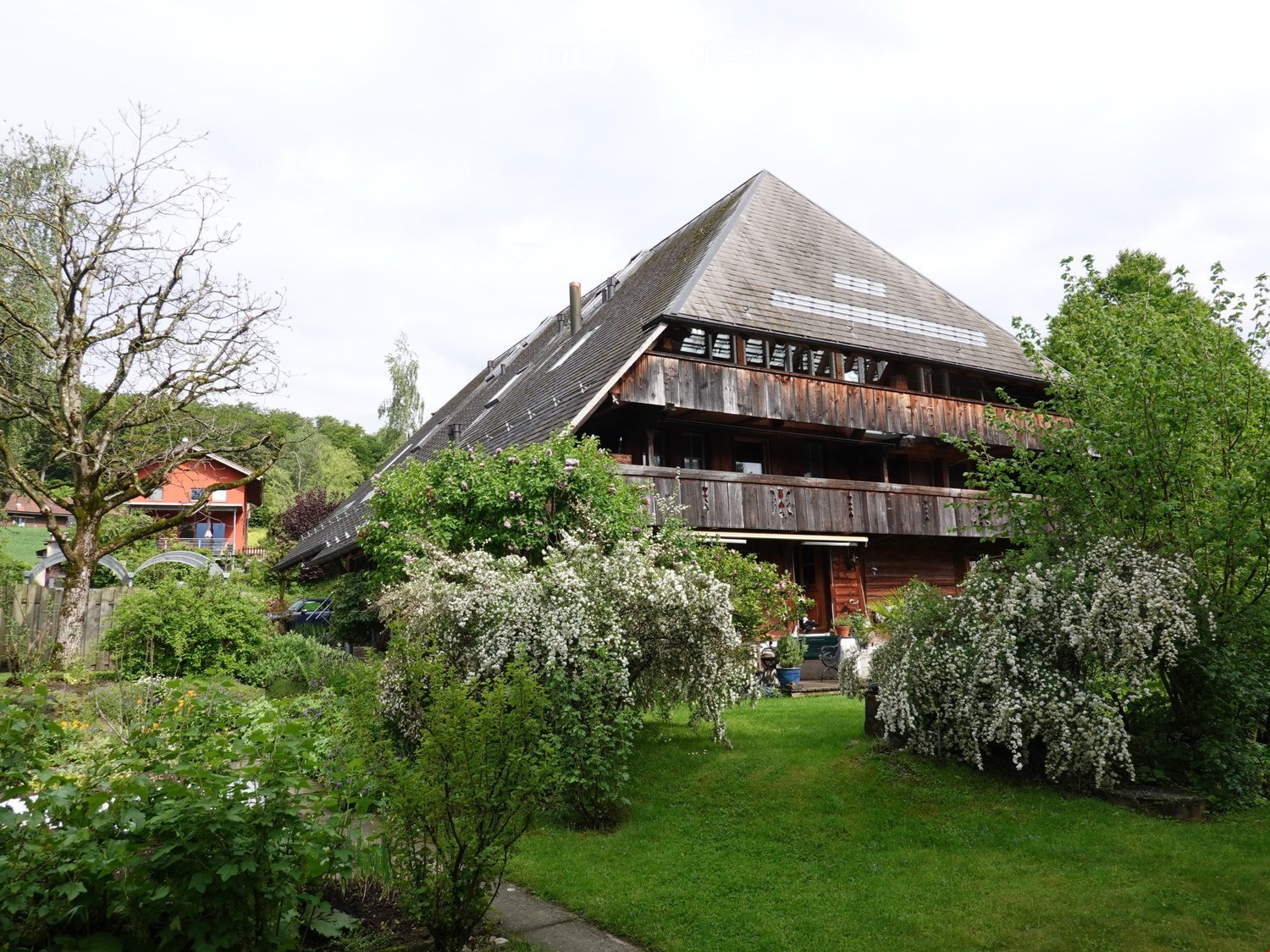 Traditional wooden house, triangular roof, wooden balcony, surrounded by garden with various plants and trees
