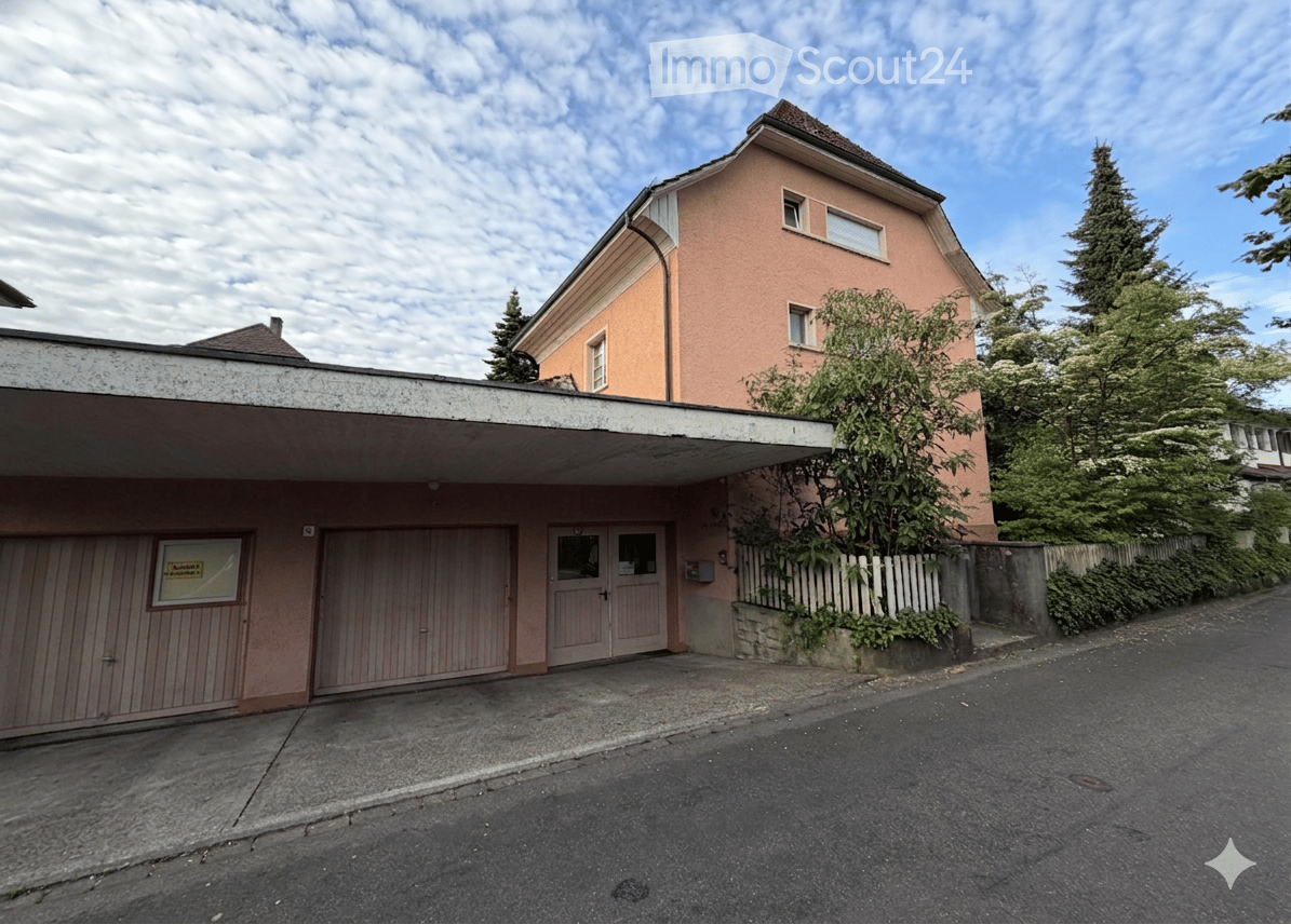 Two-story building with a garage, pink facade, and multiple windows. The entrance features a double door and a covered area.