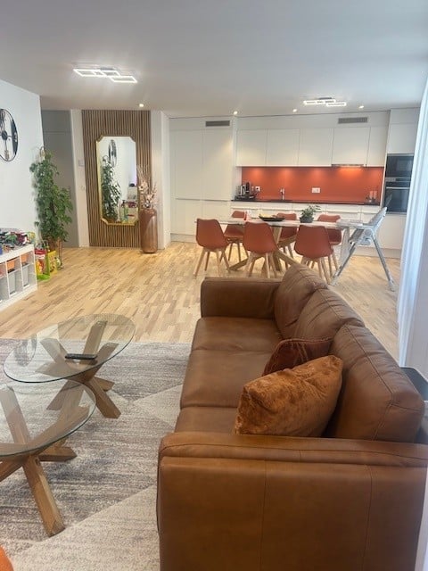 Open-plan living room with a brown leather couch, glass coffee table, and a dining area with chairs around a table.