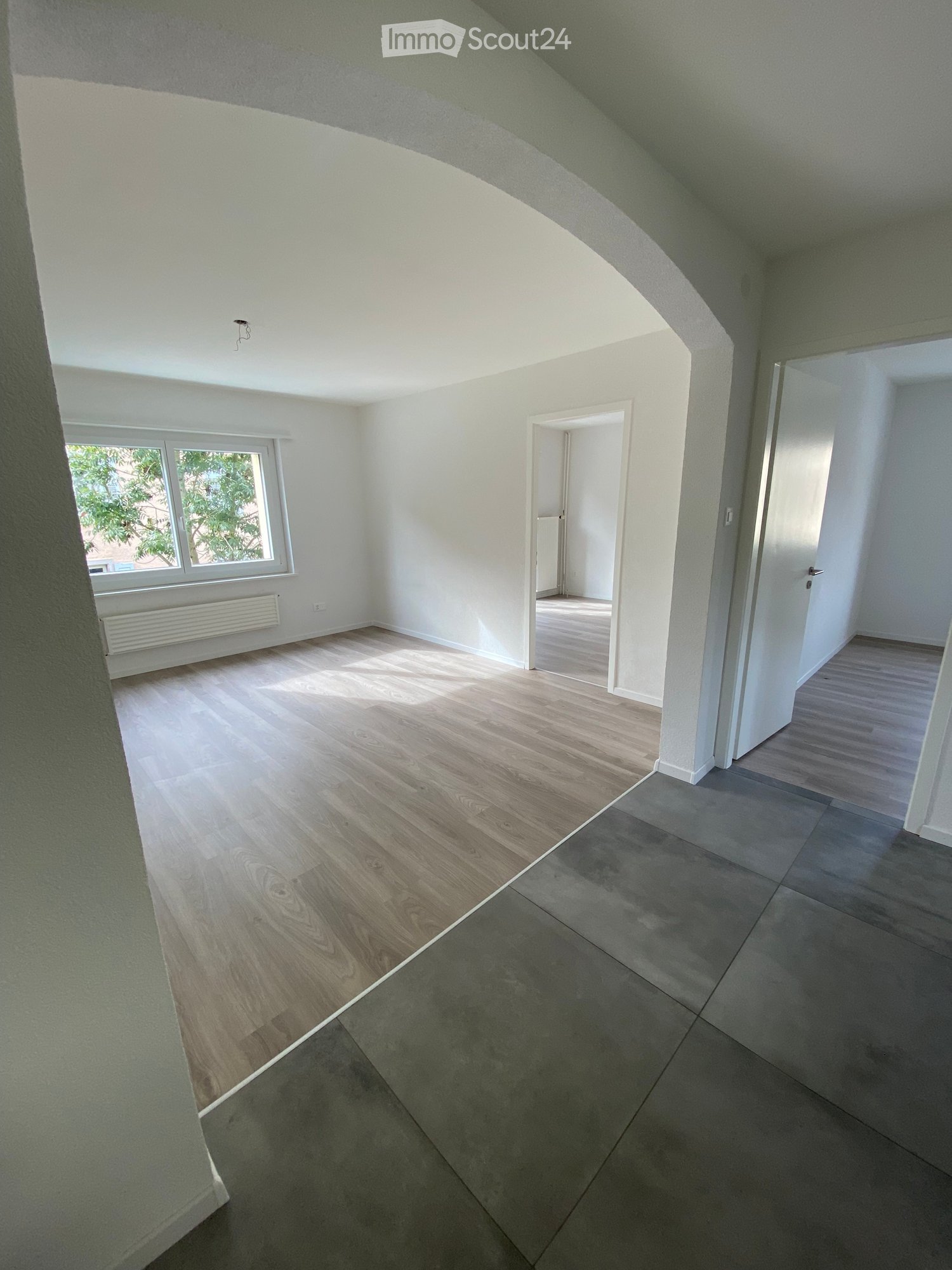 Empty room with wooden floor, white walls, window with white frame, radiator, and two doors.