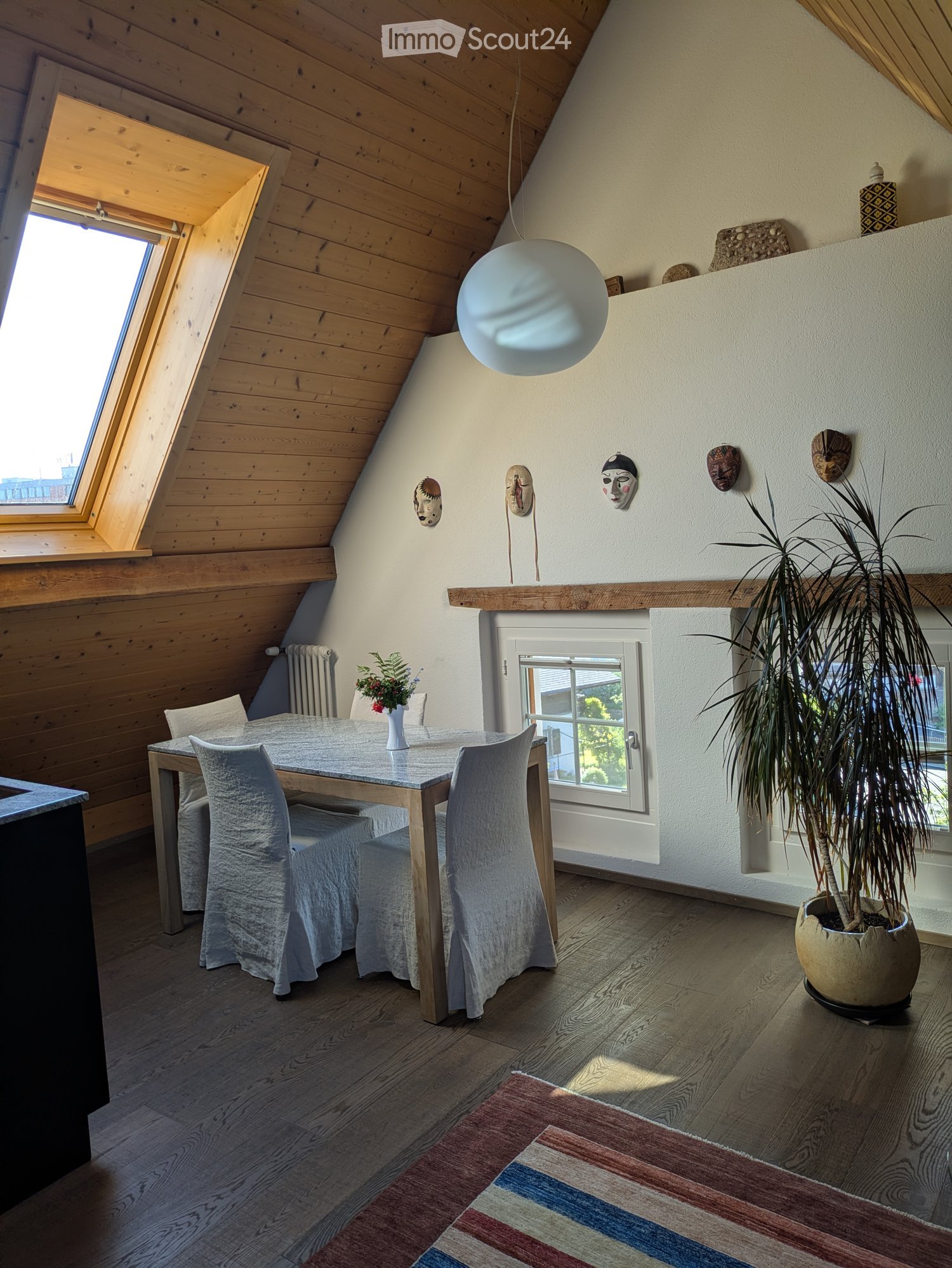 Dining area with wooden table and chairs, white covers, flower vase, wooden ceiling, wooden floor, white walls, and a potted plant.