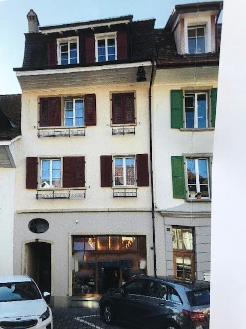 Three-story residential building with white facade, multiple windows with shutters, small balconies, and a storefront at ground level.