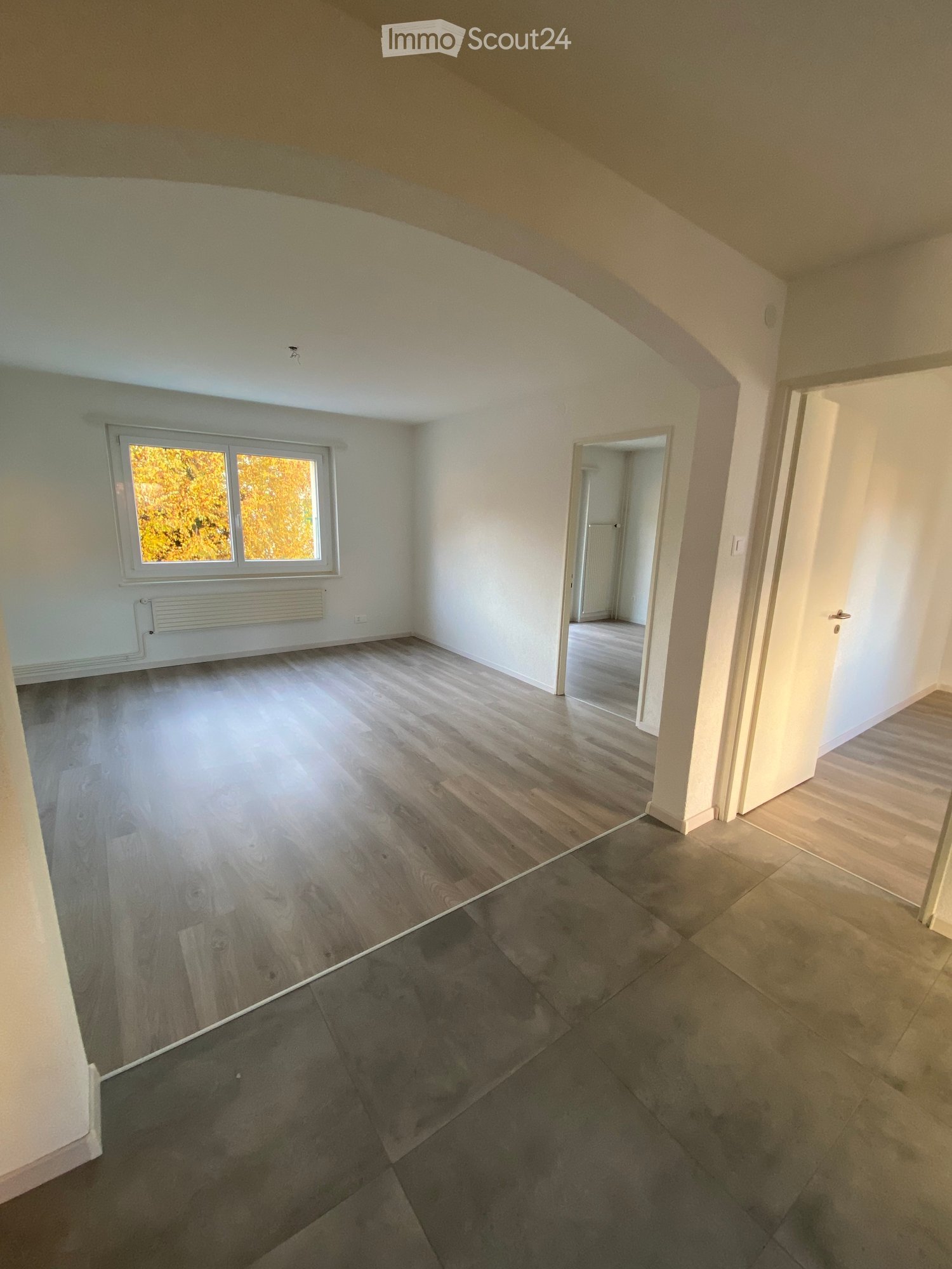 Empty room with wooden flooring, white walls, and a large window showing autumn foliage. The room has a doorway leading to another space with similar flooring.