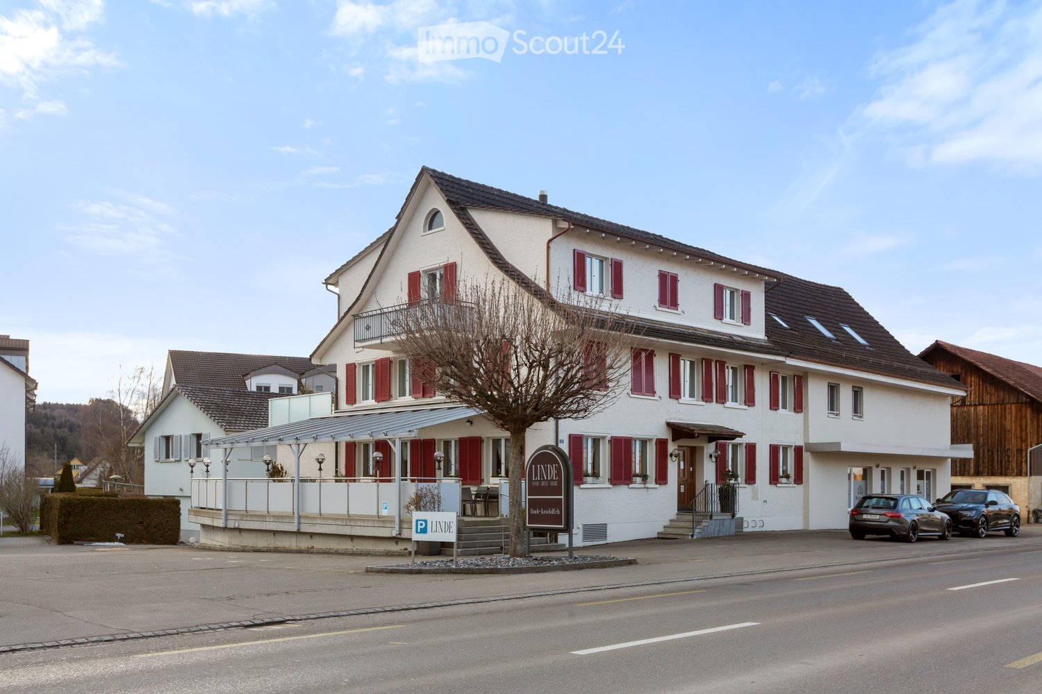 2 story white house, red shutters, brown roof, balcony, trees, parking lot, cars parked