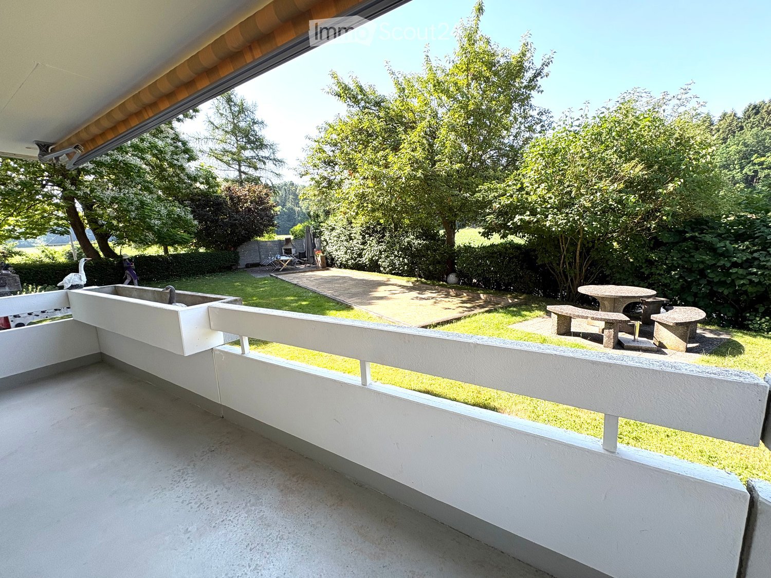 Balcony with white railing overlooking a well-maintained garden with a stone table, benches, and a sink.