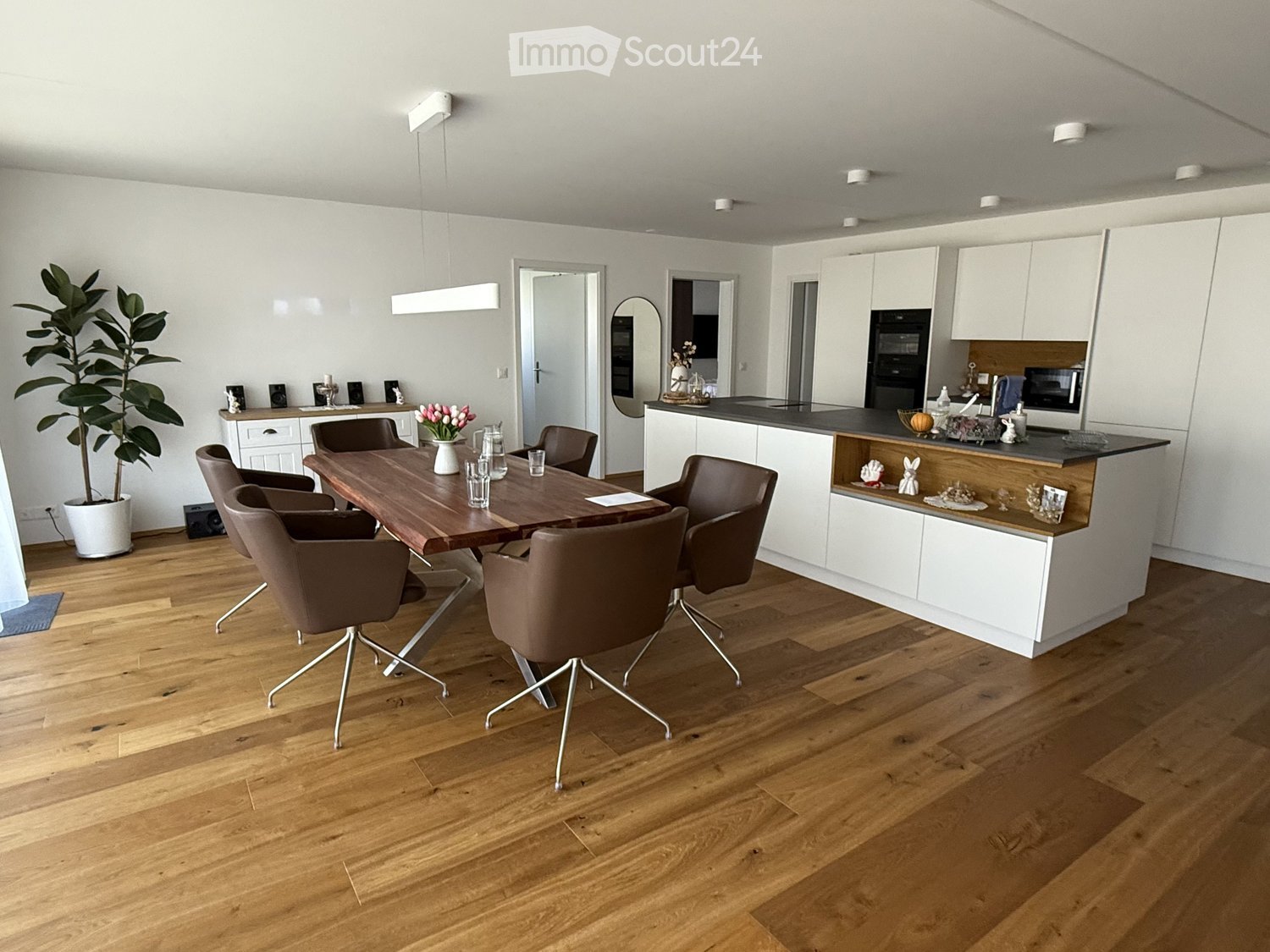 Furnished Room. Modern dining area with wooden table, brown chairs, and a white cabinet. A plant and flowers add a touch of nature. The kitchen has a counter, microwave, and oven.