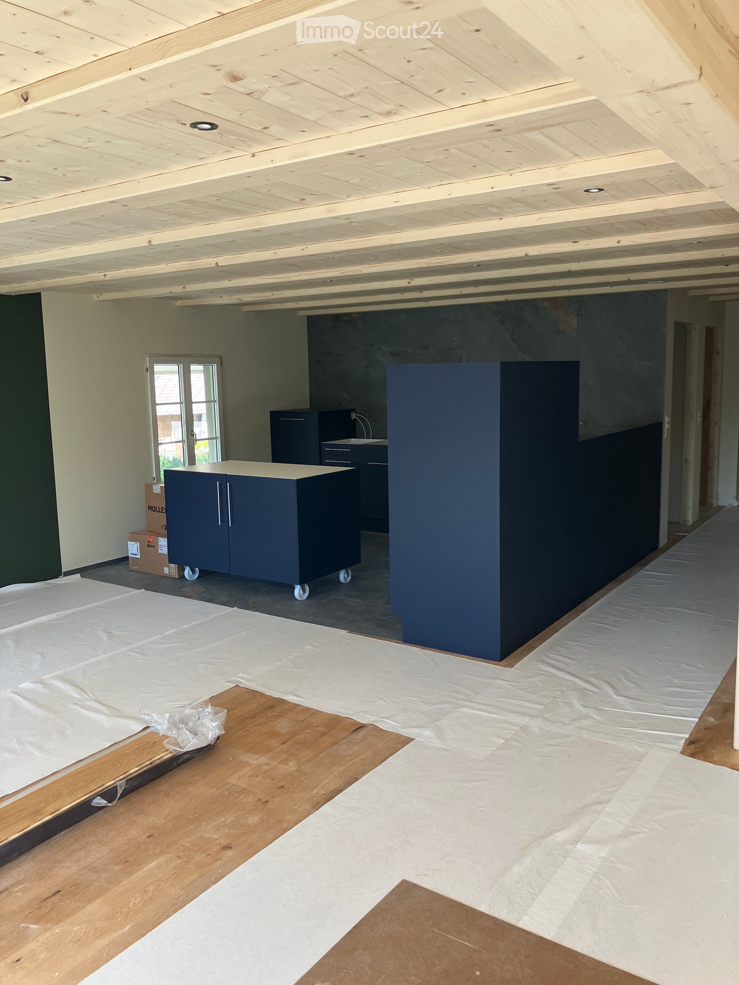 Empty room with exposed wooden ceiling beams, light-colored walls, and a mix of wooden and concrete flooring. Some plastic sheeting is on the floor, and there are a few cardboard boxes and a small wooden plank visible.