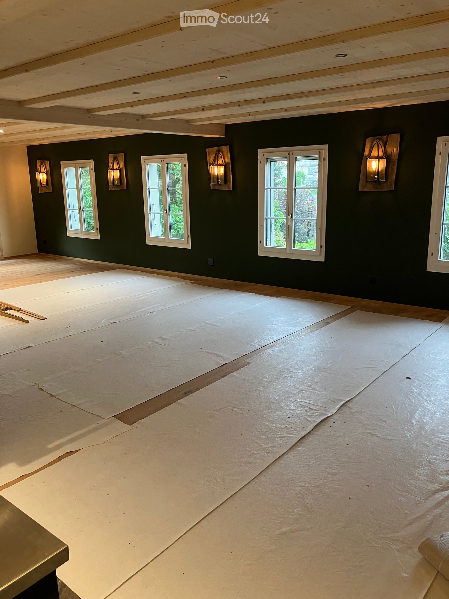 Empty room with dark green walls, wooden floor, and several windows. Exposed wooden beams on the ceiling and wall-mounted lamps are visible.