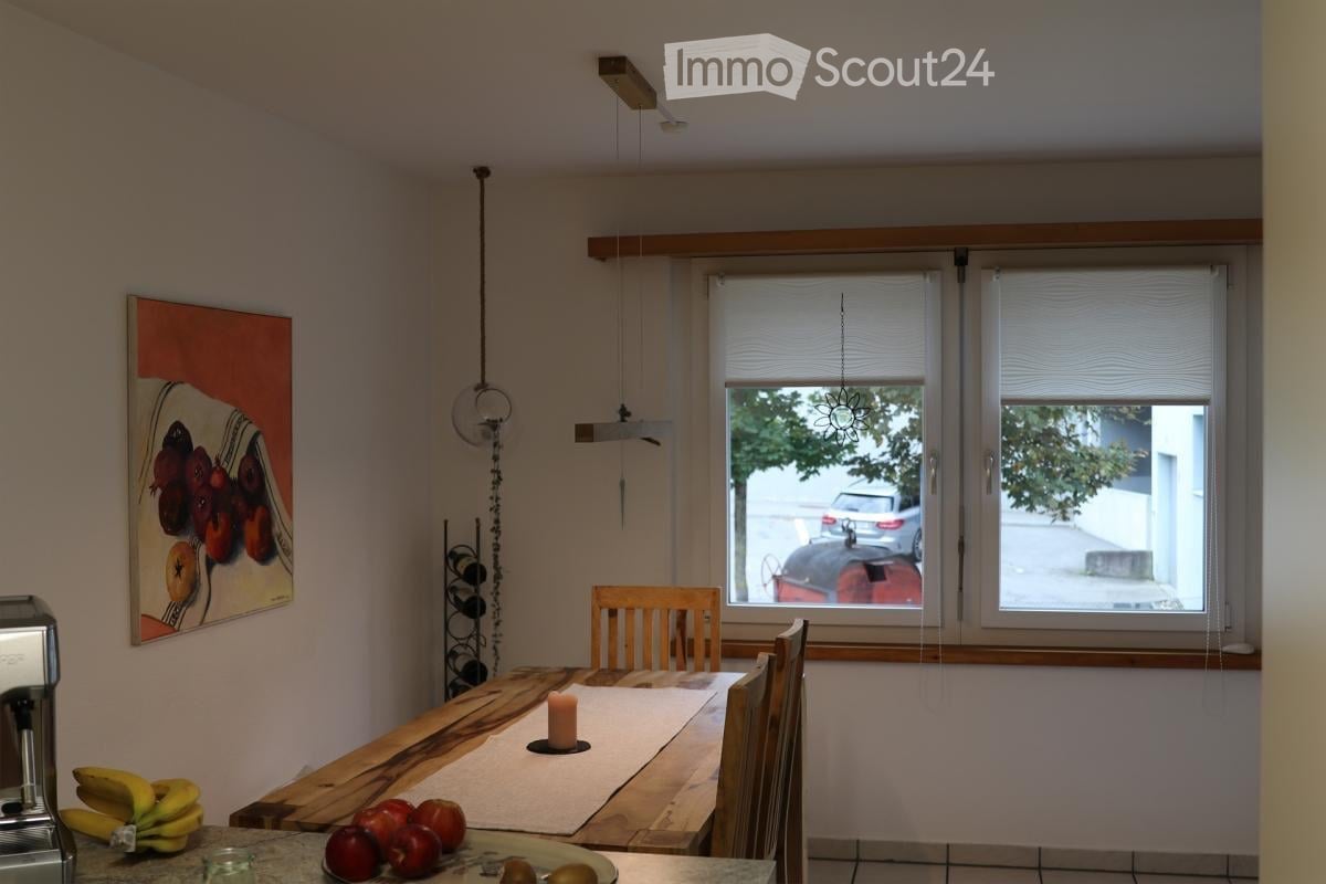 Furnished Room. Dining area with wooden table, chairs, and a candle. Fruits on the table, painting on the wall, wine rack, and lamps. Coffee machine on the counter.