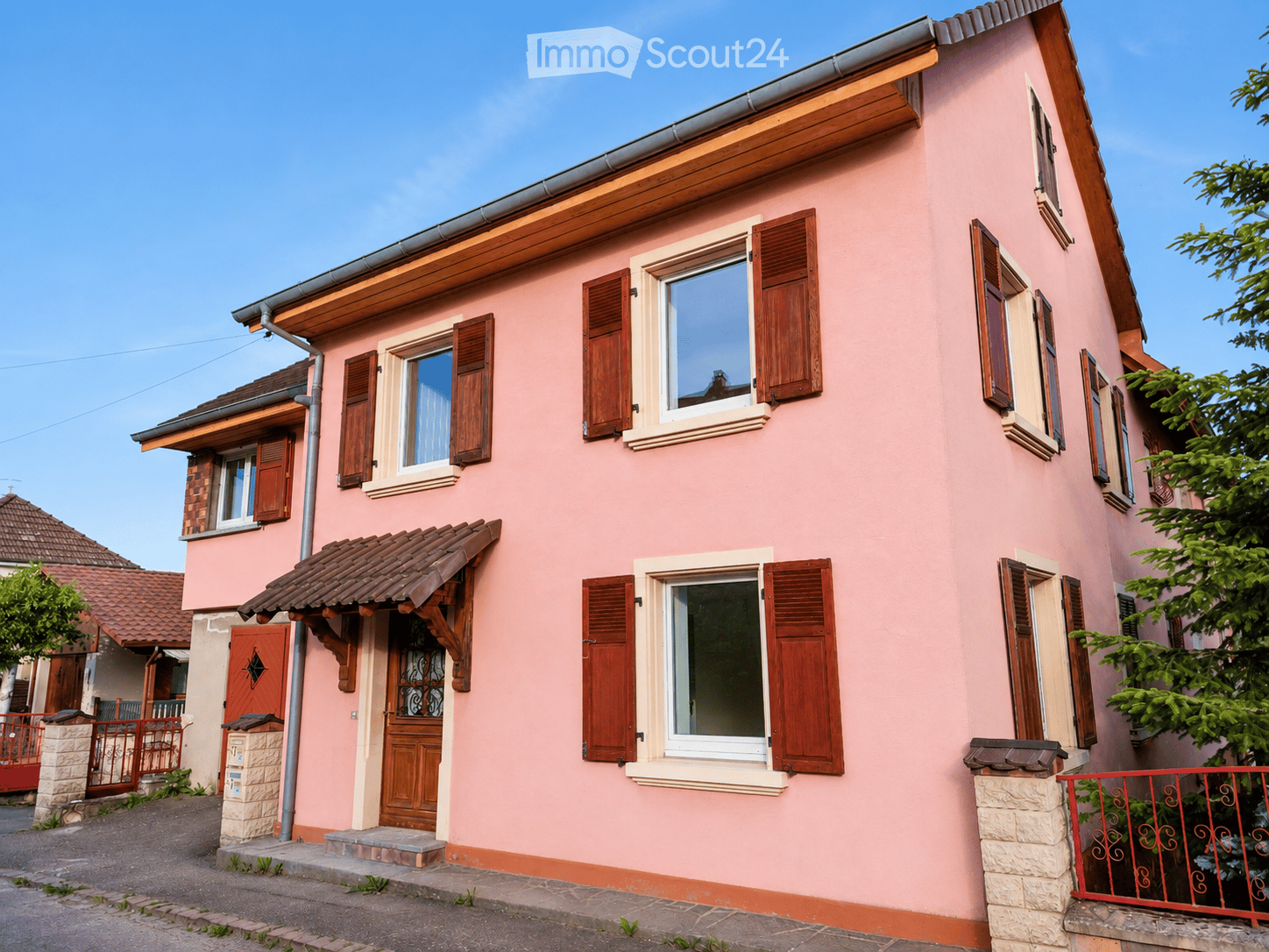 Two-story pink house with wooden shutters, multiple windows, and a tiled roof. Features a small porch with a wooden door and a stone base.