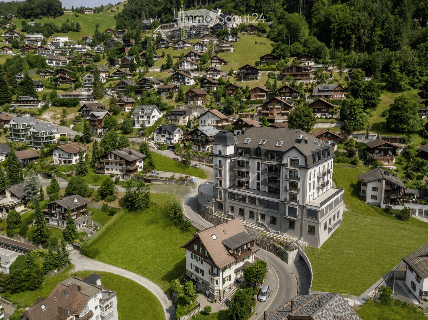 Aerial view of a residential area with multiple houses, green lawns, and trees, surrounded by hilly terrain and forested areas.