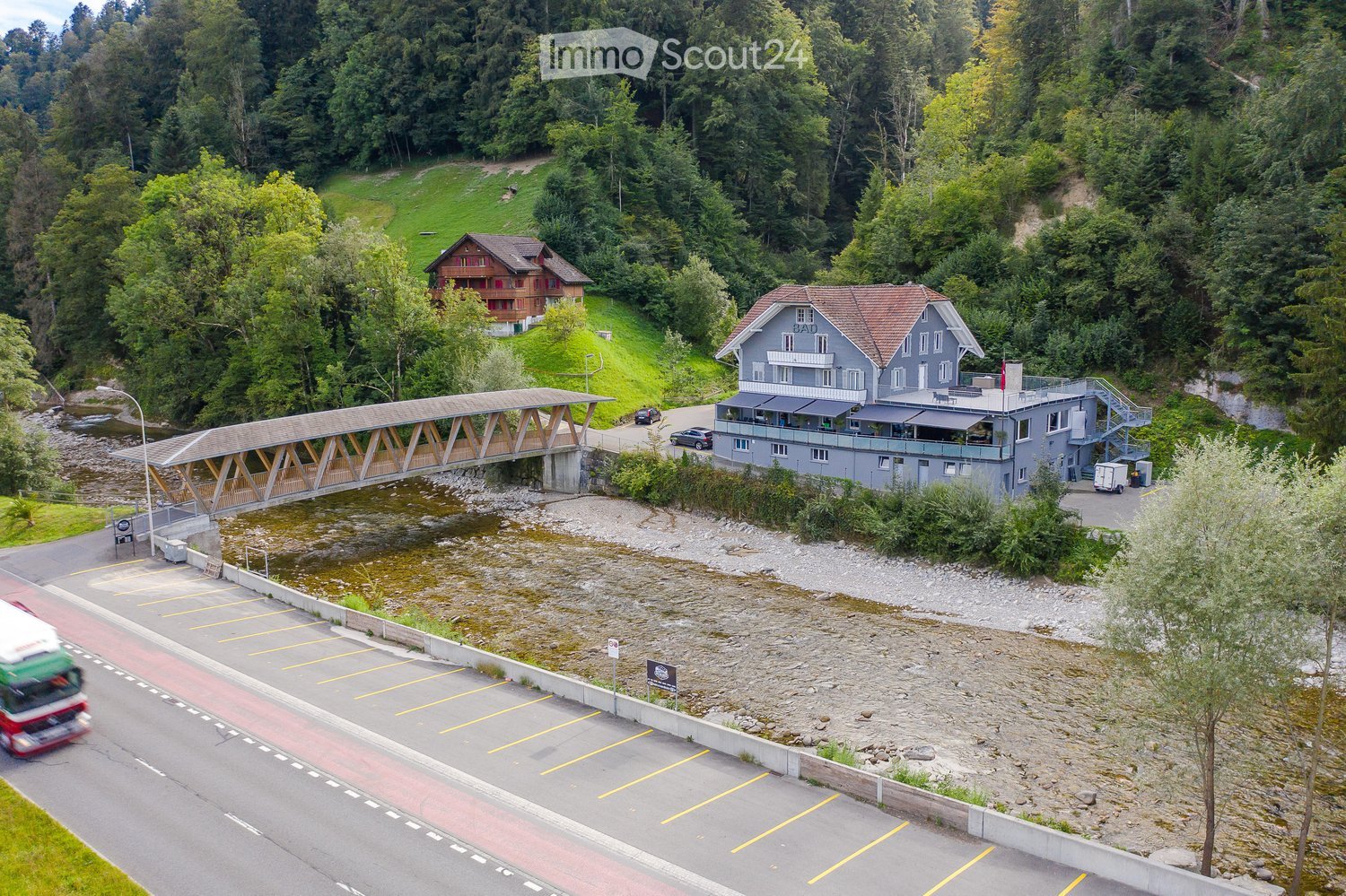 A large, modern building with a grey facade and red-tiled roof, situated near a river with a wooden bridge crossing it. The building has multiple windows and a balcony area, surrounded by lush green trees and a parking lot nearby.