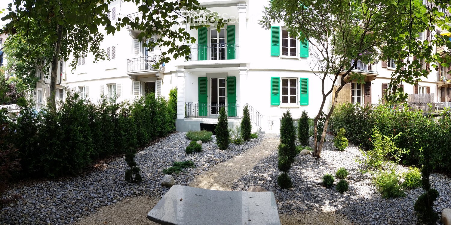 A garden area with gravel paths, shrubs, and trees in front of a white building with green shutters.