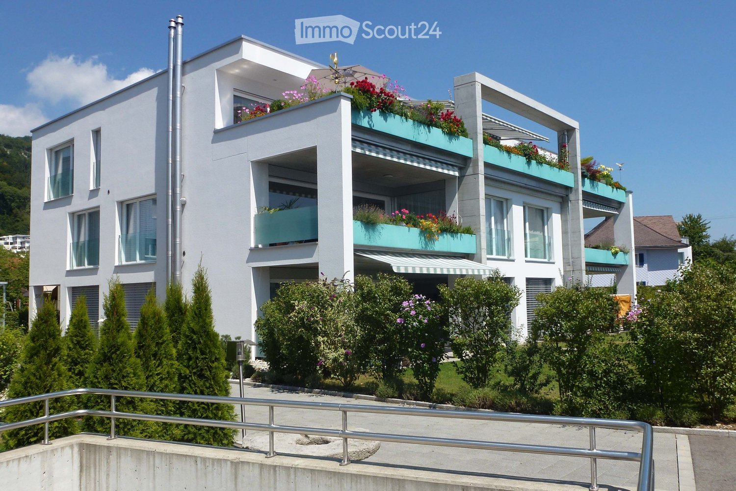 white building, 3 stories, 5 balconies, plants, trees, bushes
