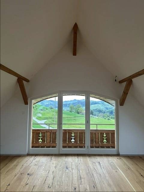 Empty room with wooden floor, white walls, and large glass doors with wooden frames. View of a green field and mountains.