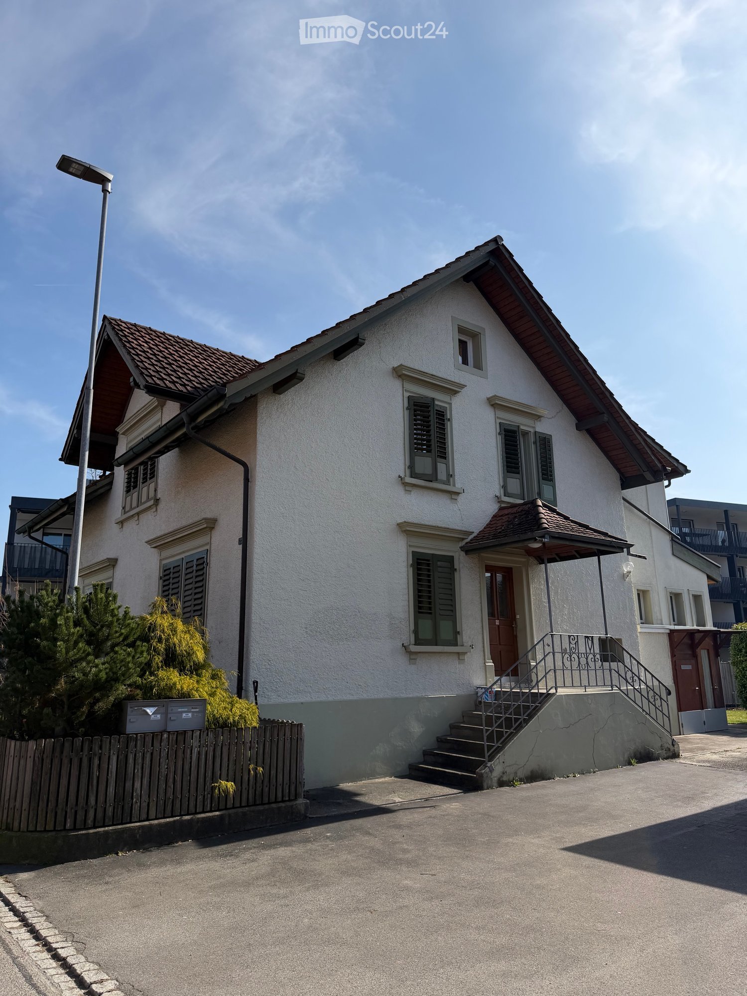 House. Two-story house with a brown roof, white walls, and green shutters. The entrance is covered by a canopy, and there are stairs leading up to the front door. A streetlight is on the left side of the house, and a wooden fence is in front of the house.