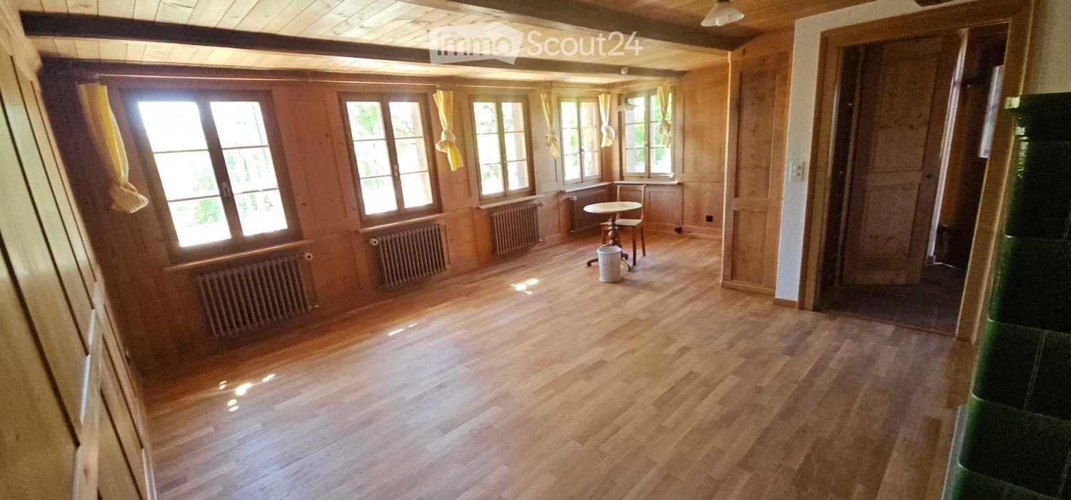Empty room with wooden flooring, wooden walls, large windows with yellow curtains, radiators, and a small round table with a chair near the windows.