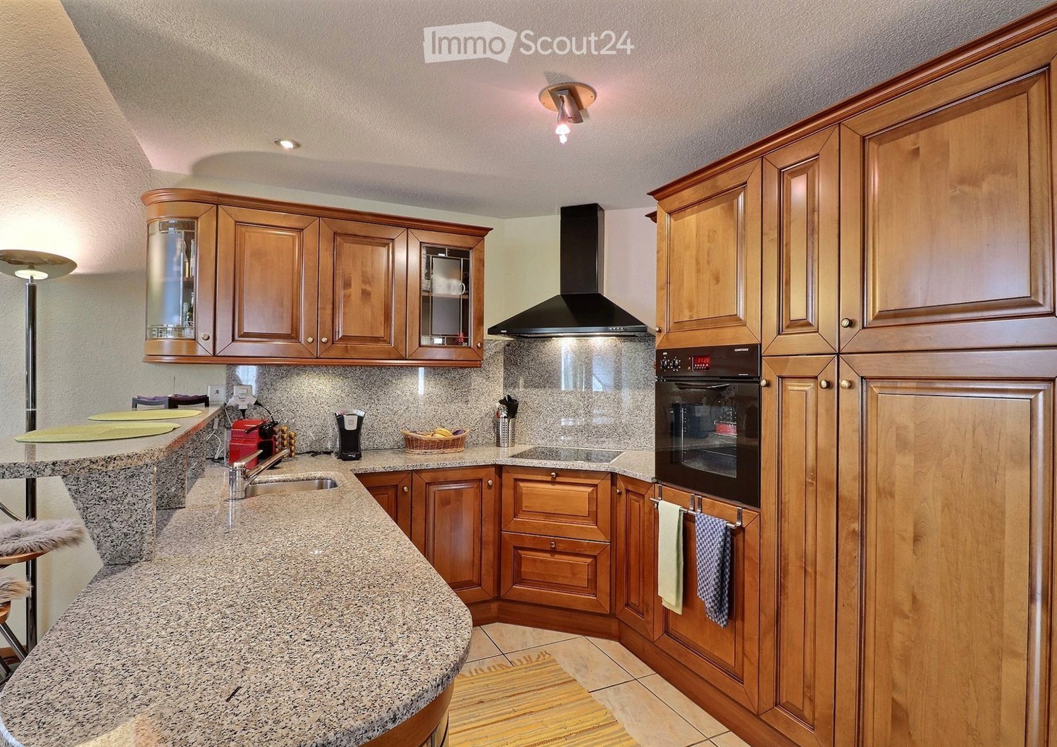 A modern kitchen with wooden cabinets, granite countertops, a stainless steel oven, range hood, and various appliances. The space includes a sink, coffee maker, and a small basket on the counter.