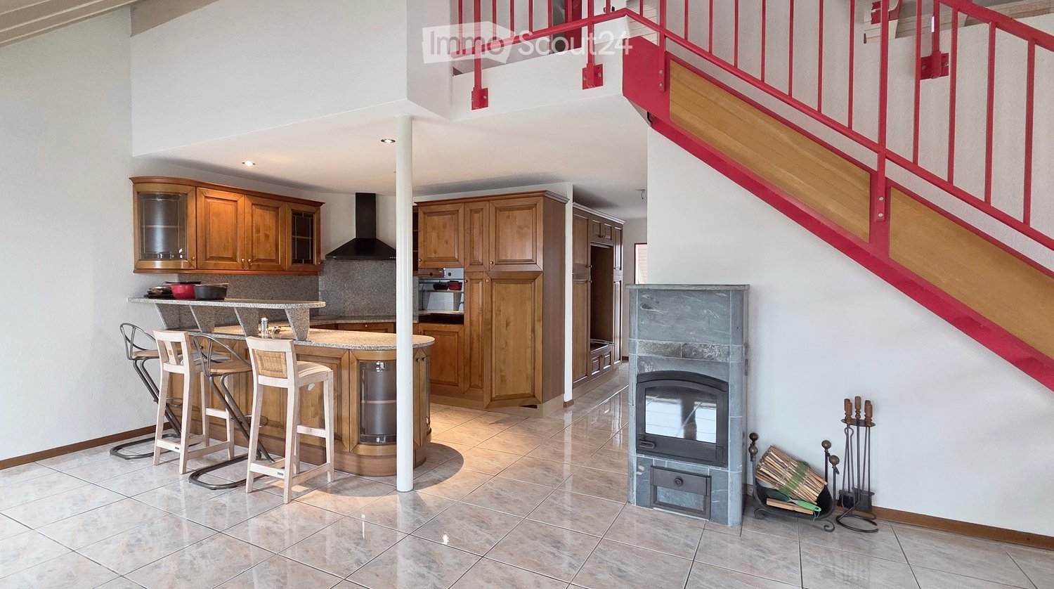 Open-plan kitchen and living area with wooden cabinets, a central island with bar stools, a modern range hood, and a fireplace with a firewood rack nearby.