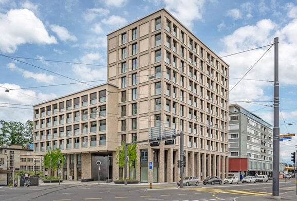 Modern multi-story building with a symmetrical facade, large windows, and a flat roof, situated on a city street with traffic and surrounding structures.