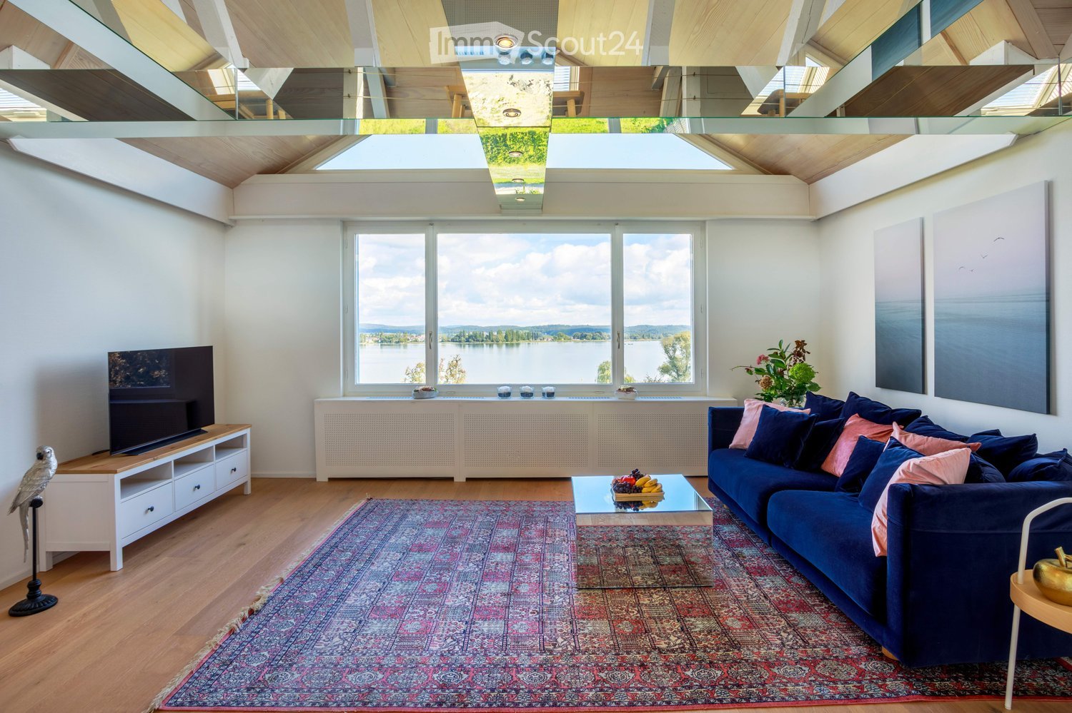 Modern living room with large windows offering a scenic view, a blue sectional sofa, a glass coffee table, a television on a wooden stand, and a decorative birdcage.