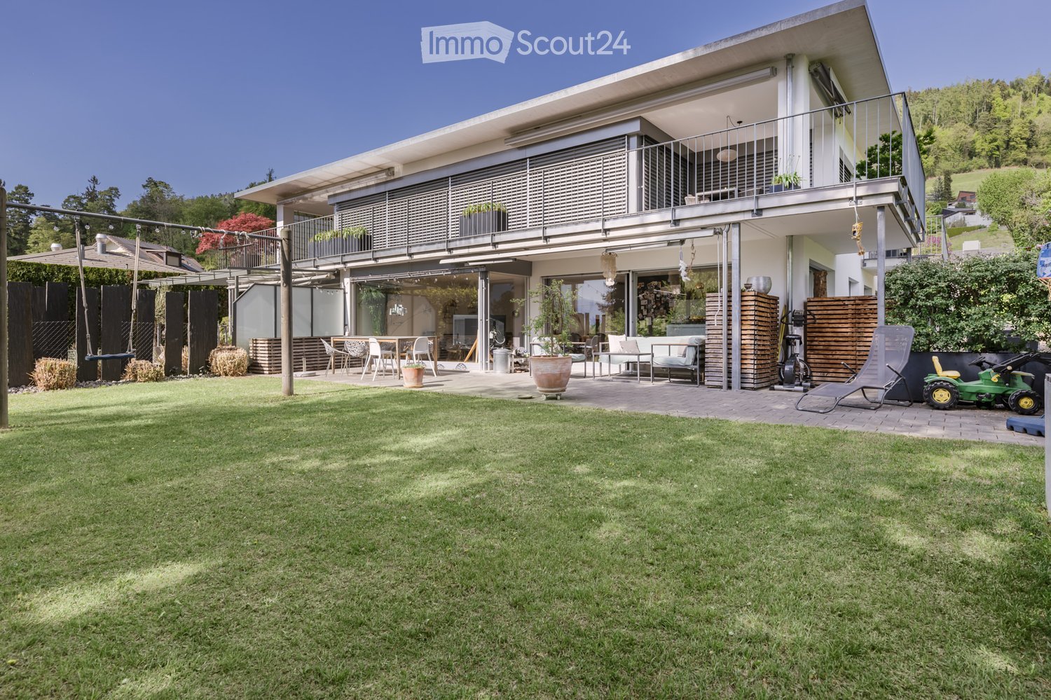 Outdoor area with a large lawn, a swing, a table with chairs, and a balcony with potted plants.