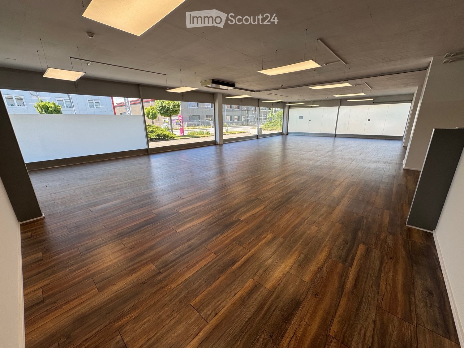 Empty room with polished wooden flooring, large windows, and modern lighting fixtures.