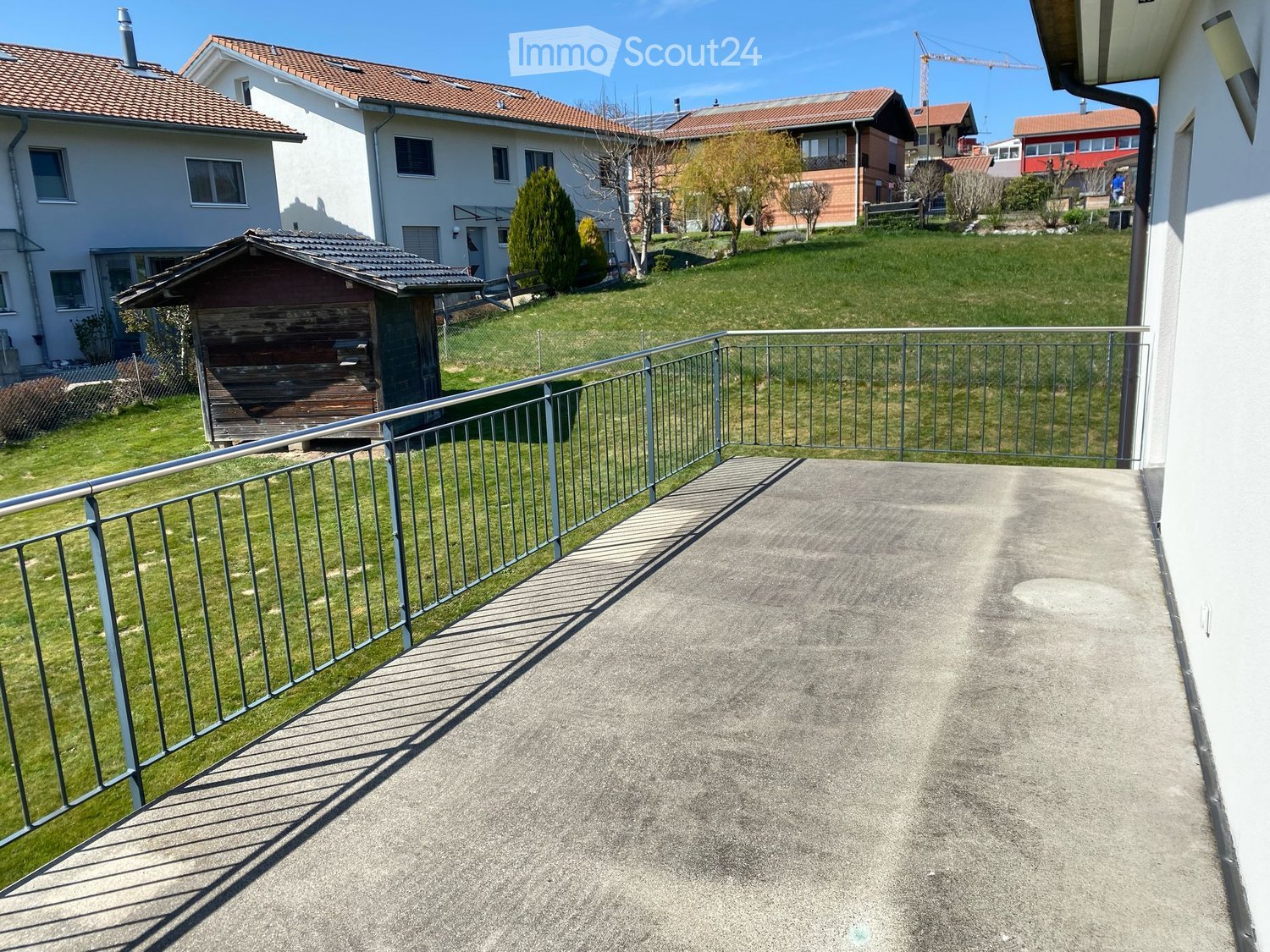 Balcony with metal railings, concrete floor, overlooking a grassy area with a small wooden shed