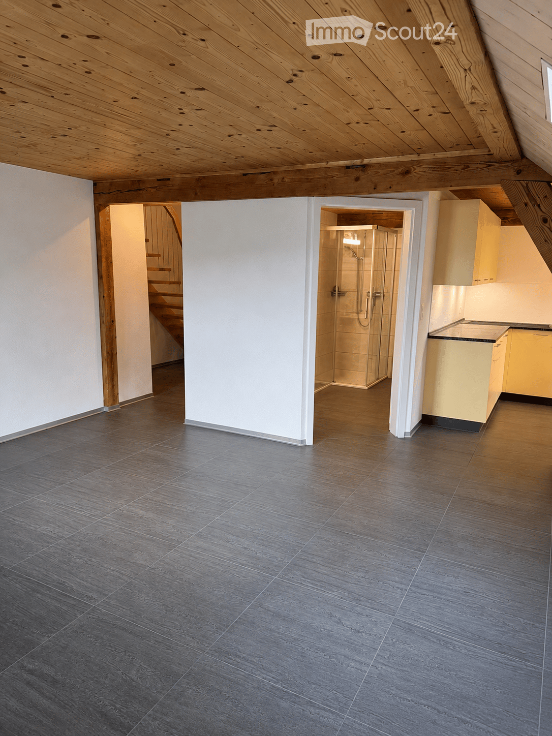 An empty room with a wooden ceiling, grey tiled floor, and visible doorways leading to a staircase and a bathroom area with a glass shower enclosure.