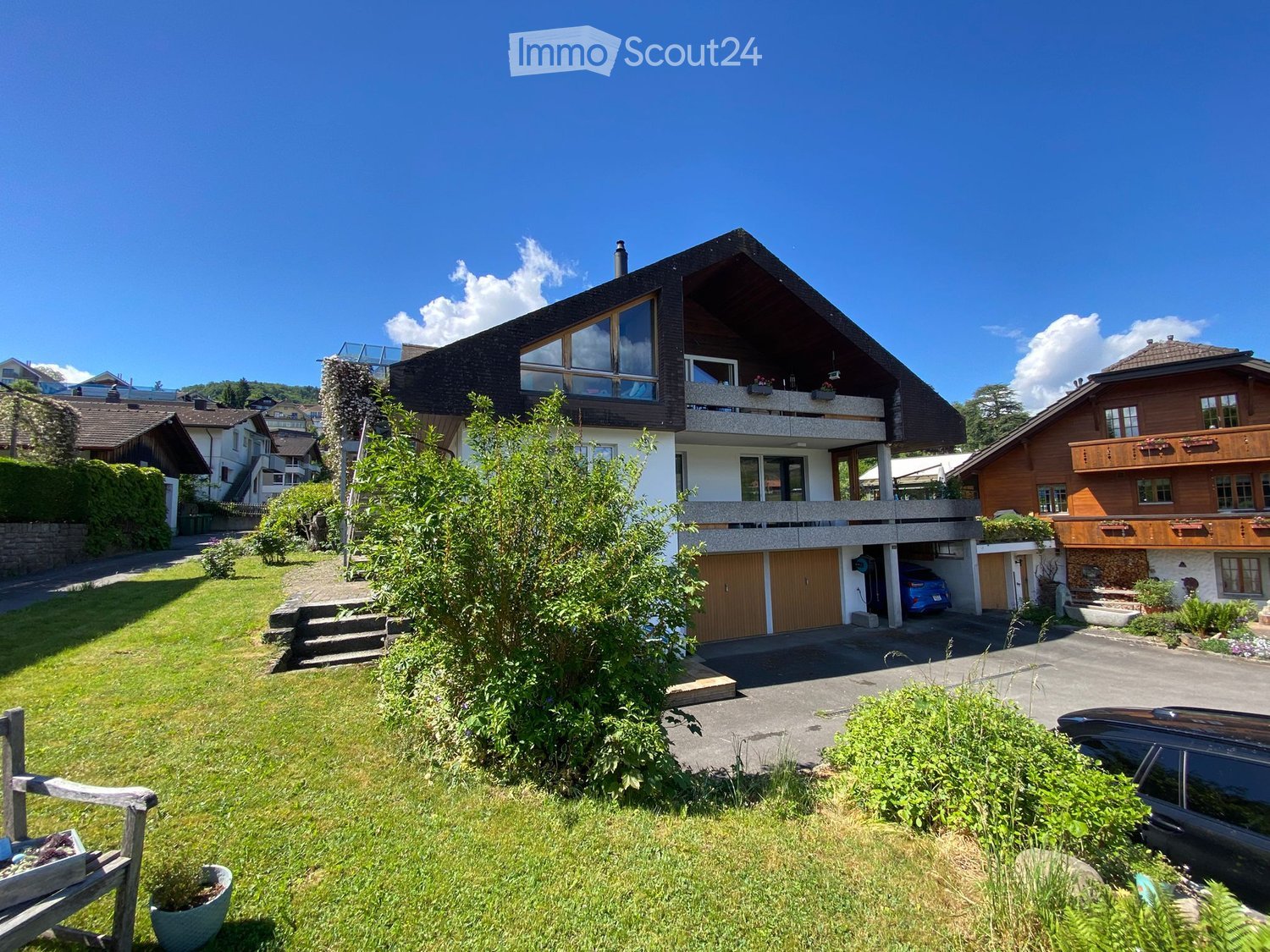 House. A two-story house with a modern design, featuring a large balcony on the upper level, a garage, and a well-maintained garden with a wooden bench and potted plants.