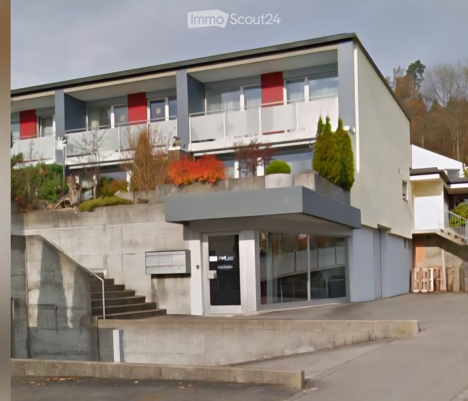 Modern two-story building with white and grey exterior, large windows, balconies, and landscaped greenery.