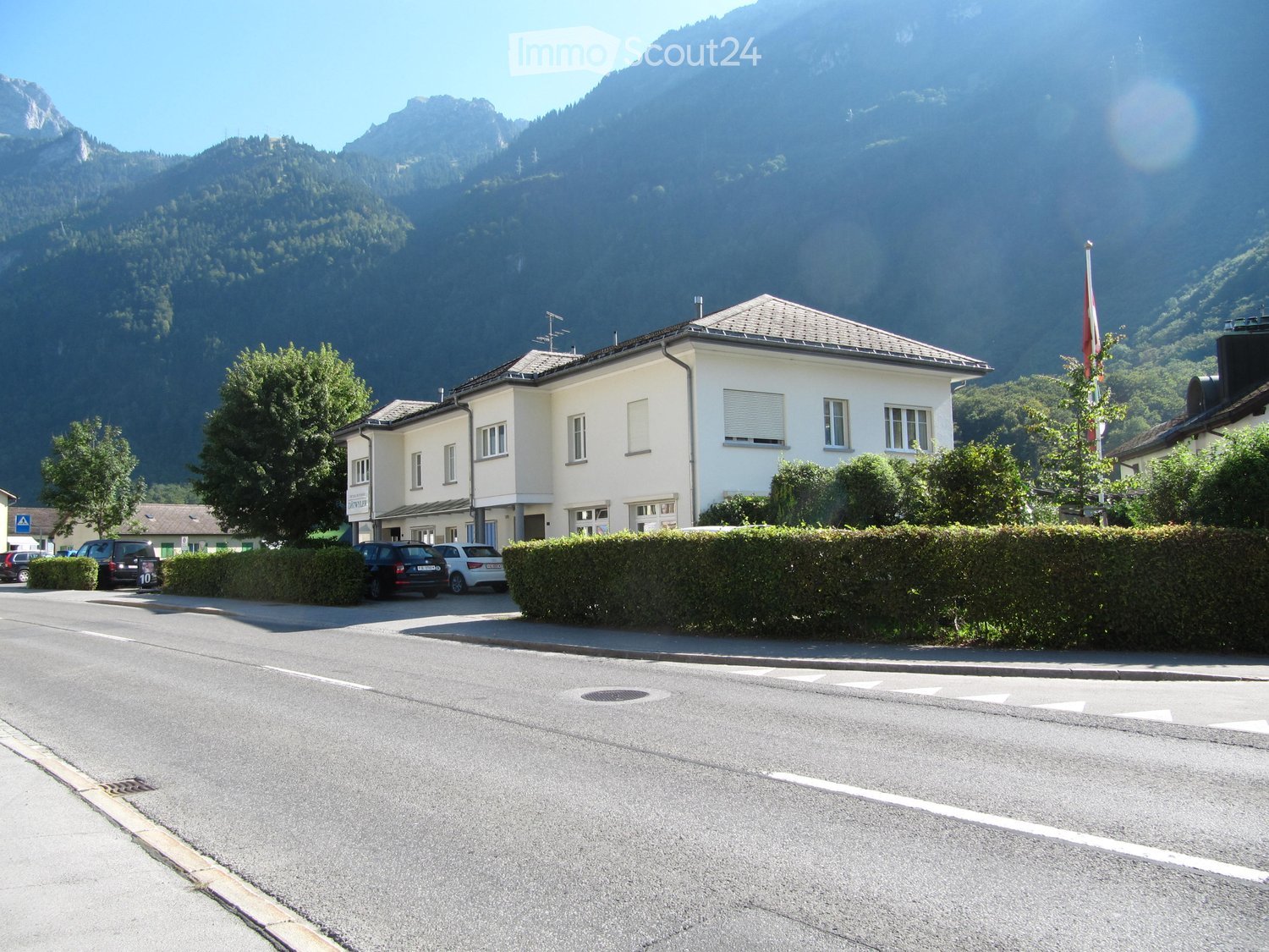 A two-story white building with multiple windows and a flagpole, situated on a street with parked cars and surrounded by greenery and mountains.
