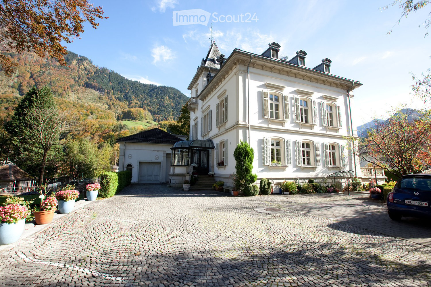 House. A large, elegant white mansion with multiple stories, classic architectural details, and a well-maintained garden. The driveway is paved with cobblestones, and there are potted plants and flowers near the entrance.