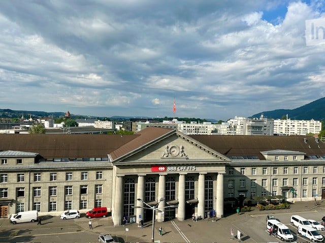 Large building with many windows, flag on top, parking lot in front, city skyline in background