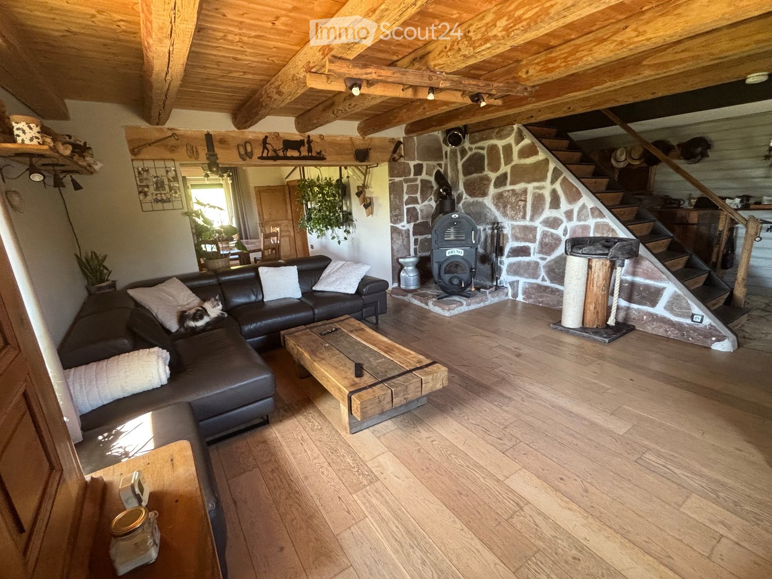 Living Room. The living room features a dark leather sectional sofa with white cushions, a wooden coffee table, a wooden staircase leading to an upper level, exposed wooden beams on the ceiling, and a stone fireplace with a wood-burning stove. The room ha