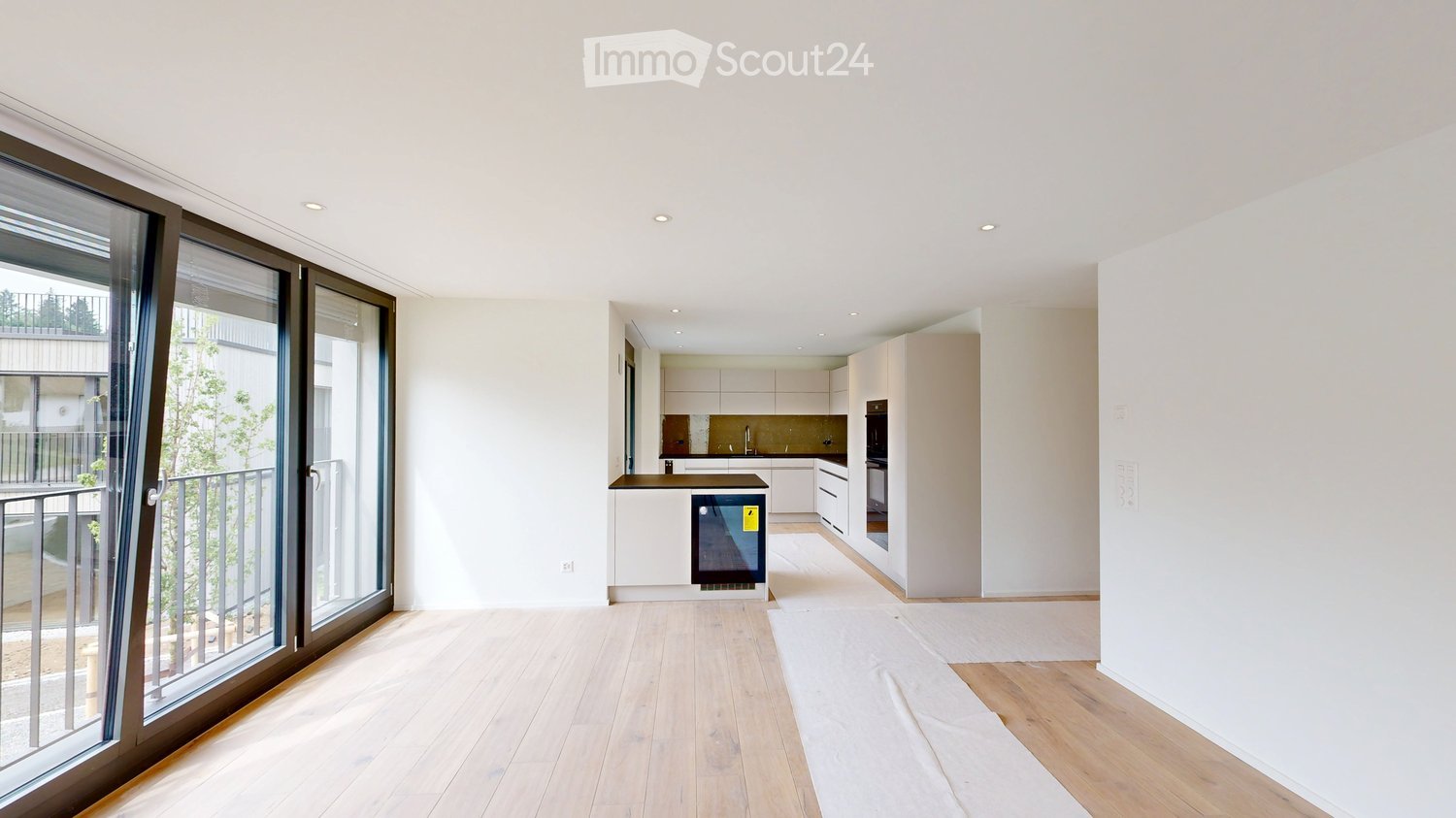 Furnished Room. Open-plan living and kitchen area with light wooden flooring, white walls, and large sliding glass doors leading to a balcony. The kitchen features modern white cabinets, a central island, and built-in appliances.