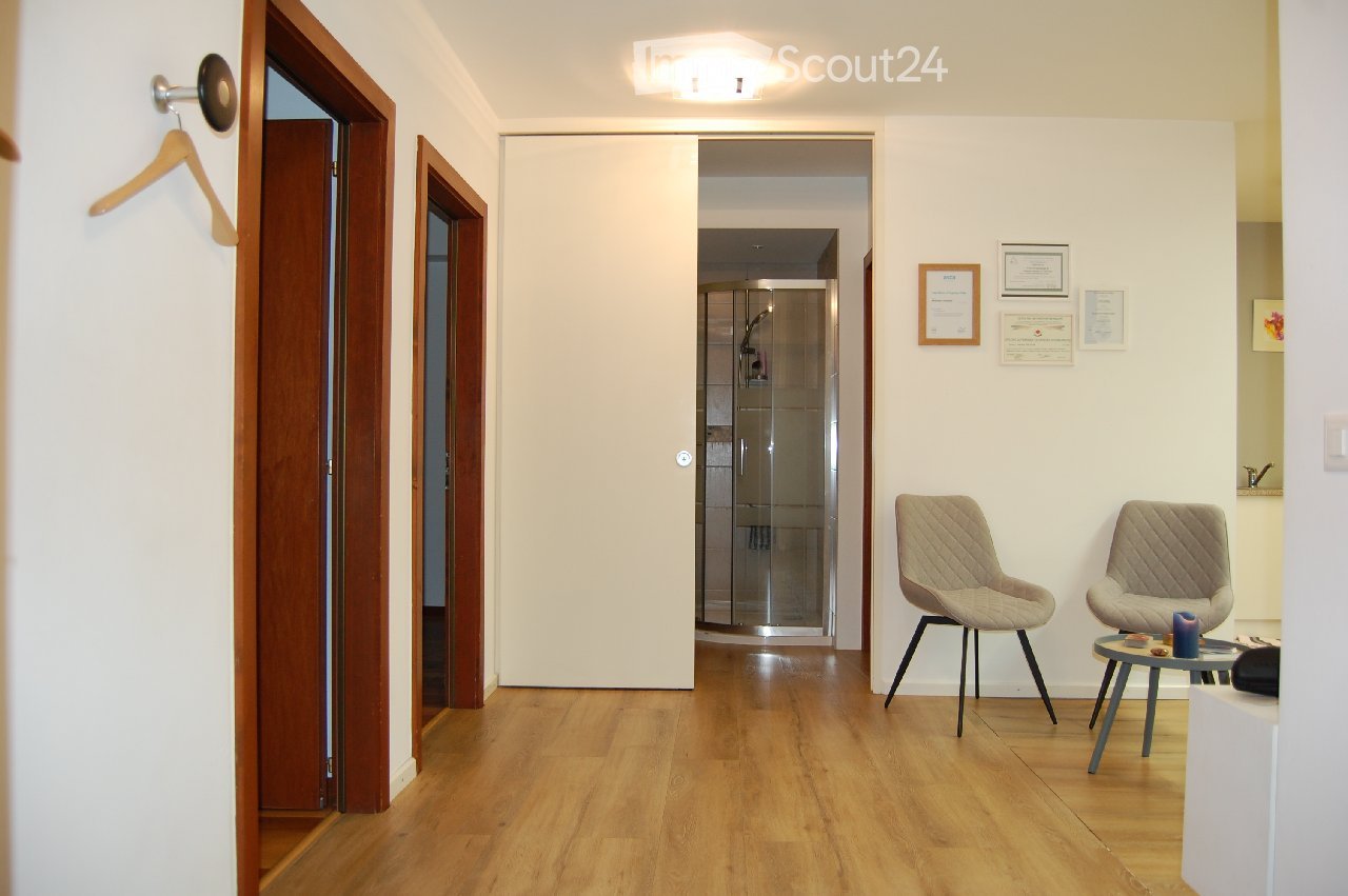Furnished Room. A modern, minimalistic hallway with wooden flooring, white walls, and a glass shower enclosure visible in the background. Two beige chairs and a small round table are placed near the wall, with framed certificates and a small decorative it