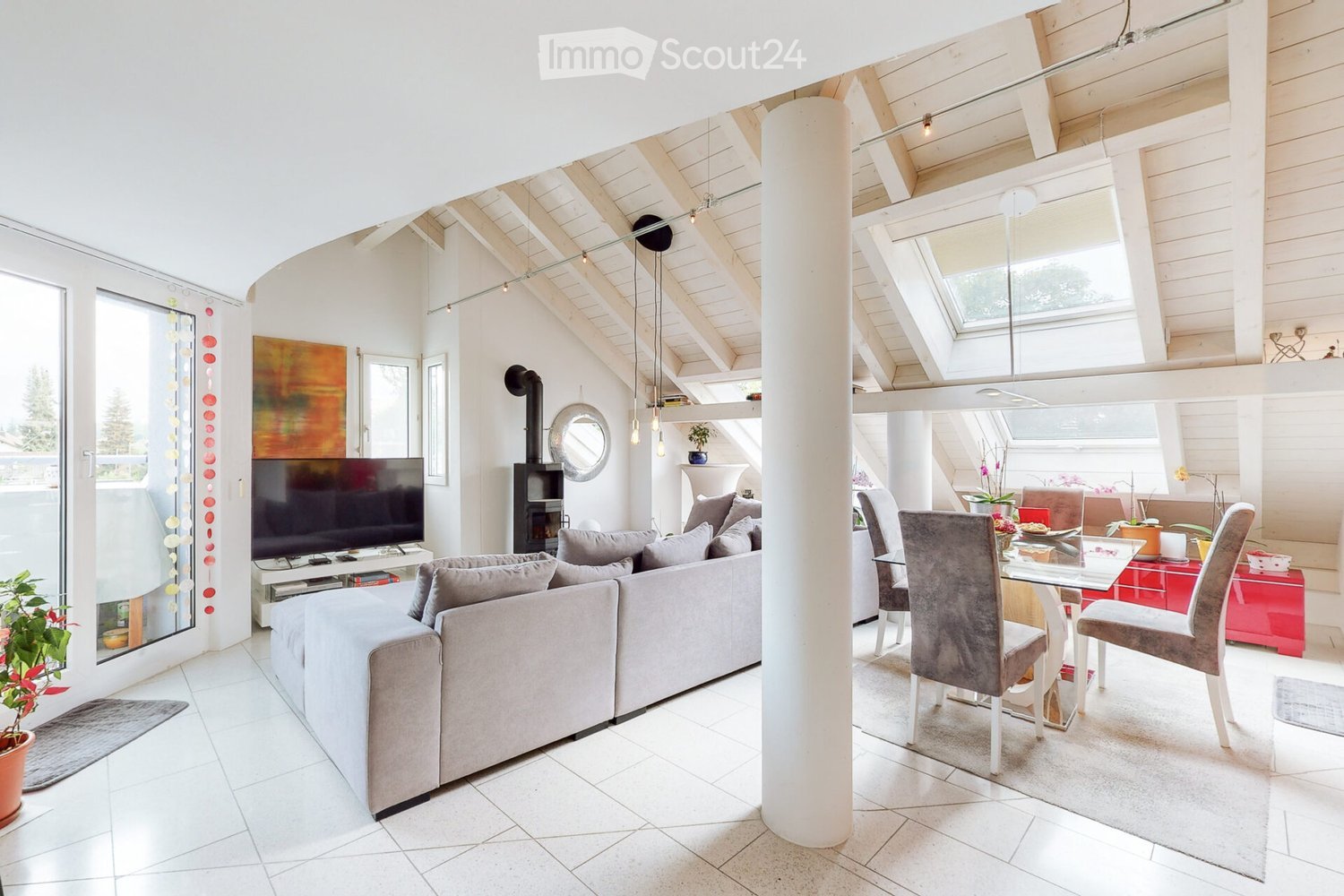 Furnished Room. Bright living and dining area with exposed wooden beams, white walls, and large windows. Features a grey sectional sofa, coffee table, flat-screen TV, fireplace, dining table with chairs, and potted plants.