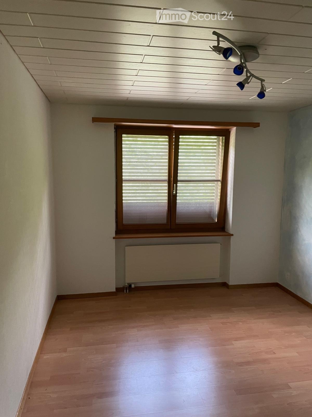 Empty room with wooden flooring, white walls, a window with blinds, a radiator, and a ceiling light fixture.