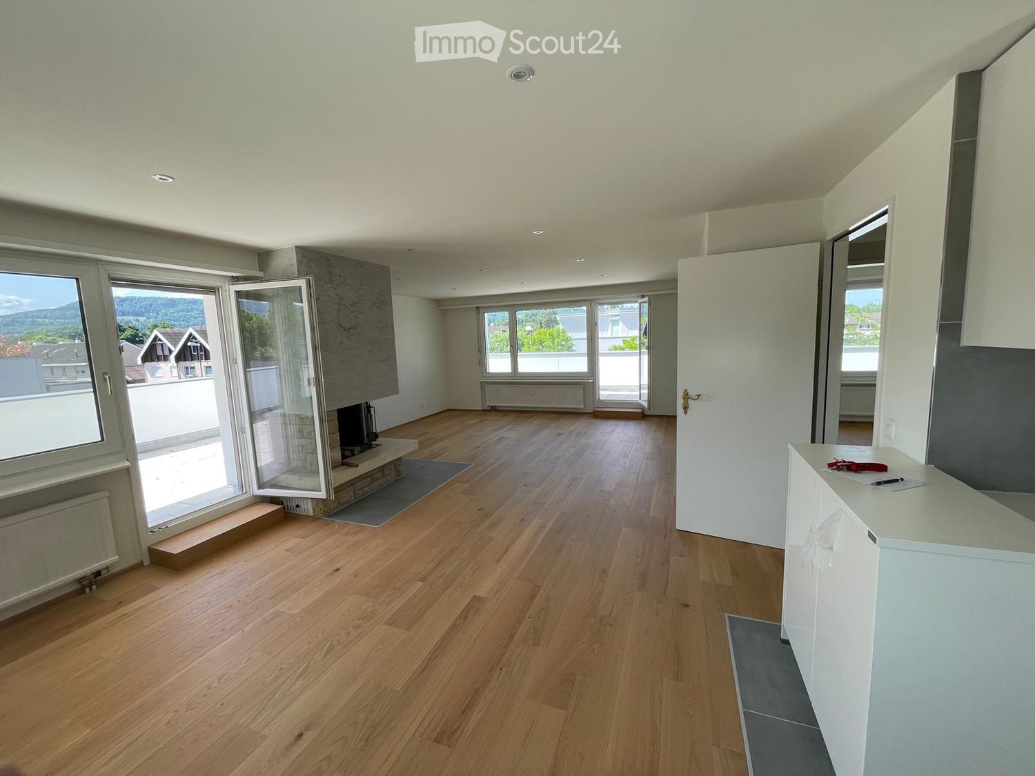 Empty room with wooden flooring, large windows offering a scenic view, and a fireplace area.