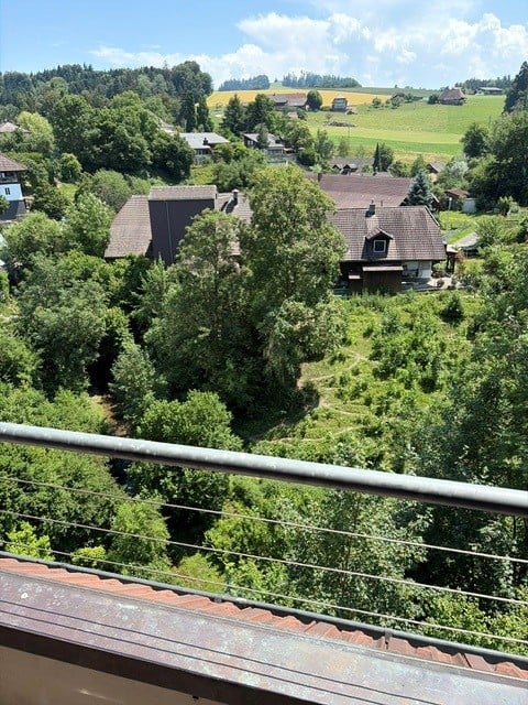 Balcony overlooking a rural landscape with scattered houses, trees, and open fields under a partly cloudy sky.