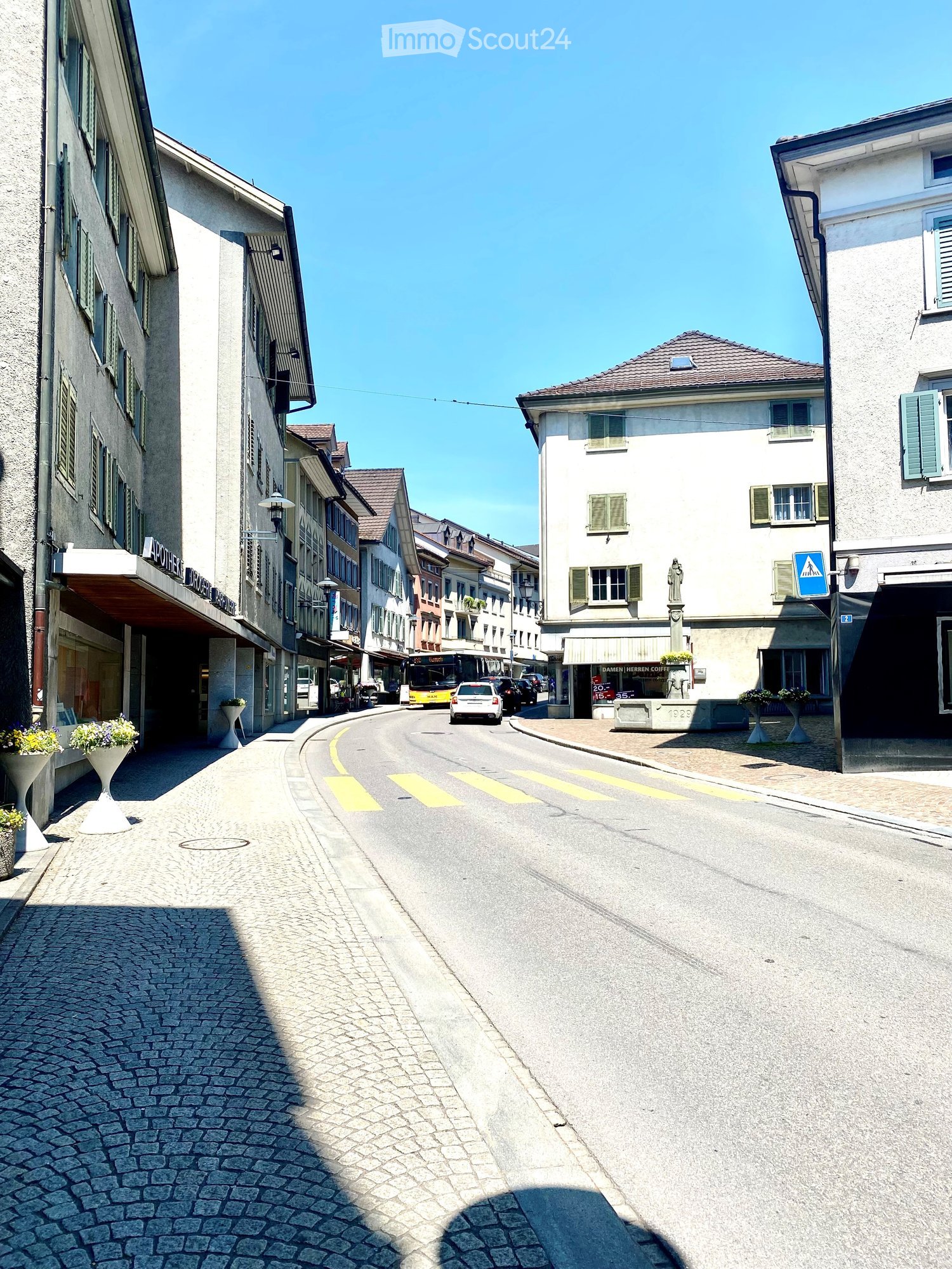 A quiet, tree-lined street in a European town with classic multi-story buildings, cobblestone pavement, and a few parked cars.