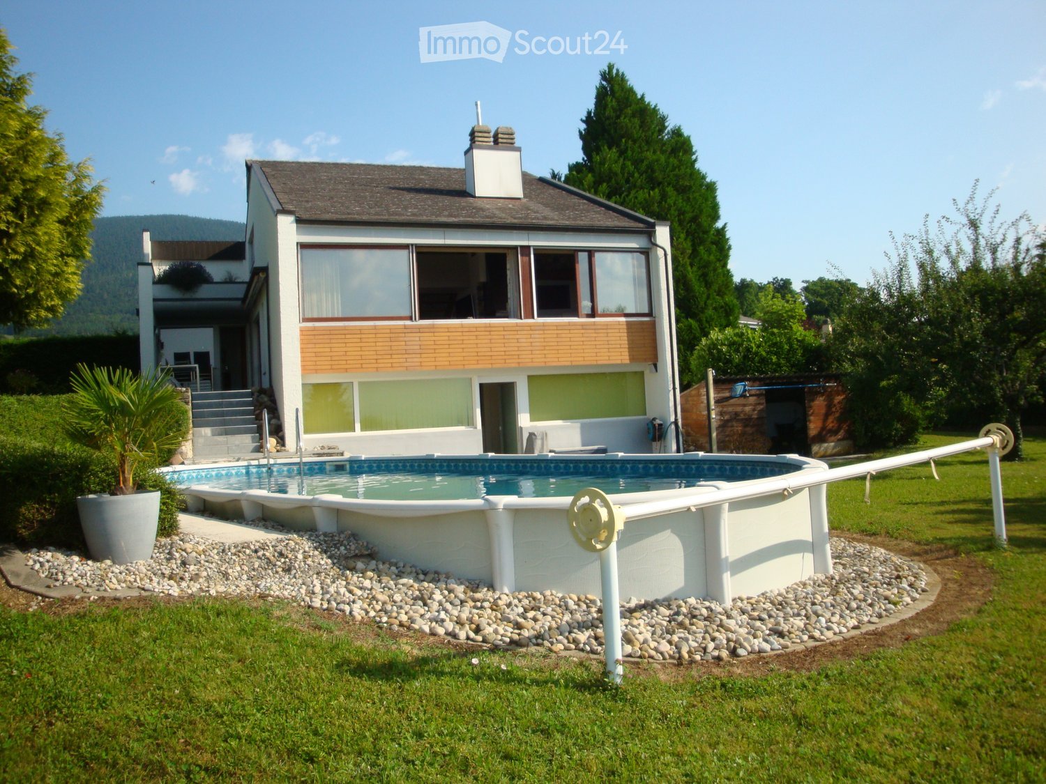 Modern house with swimming pool, large windows, brown roof, white walls, green lawn, trees, mountains in the background