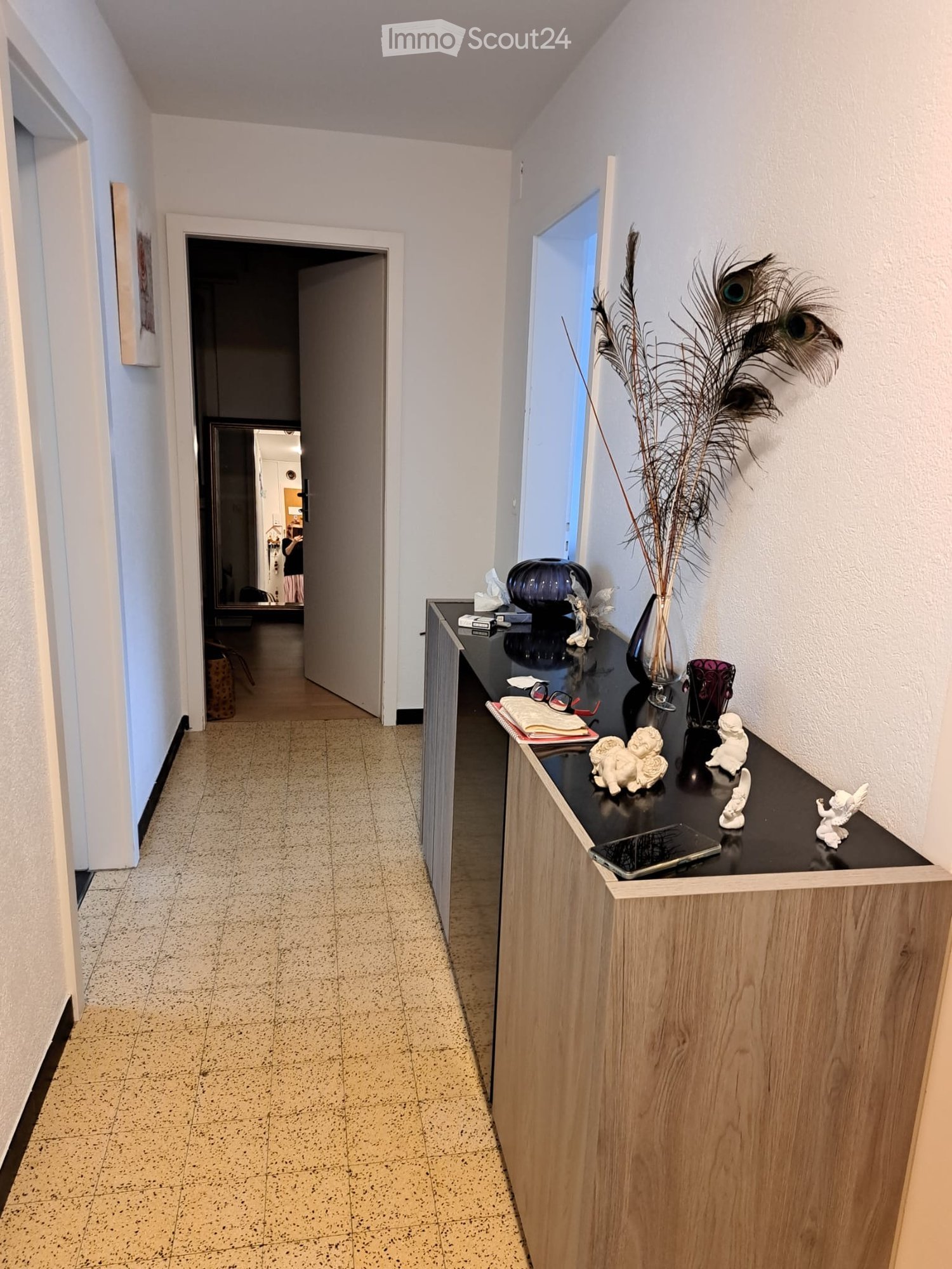 Empty hallway with a wooden floor, white walls, and a wooden cabinet with a black top. The cabinet has a vase with feathers, a purple vase, a book, a cell phone, and other items on top. There is a picture frame on the wall and an open door leading to anot