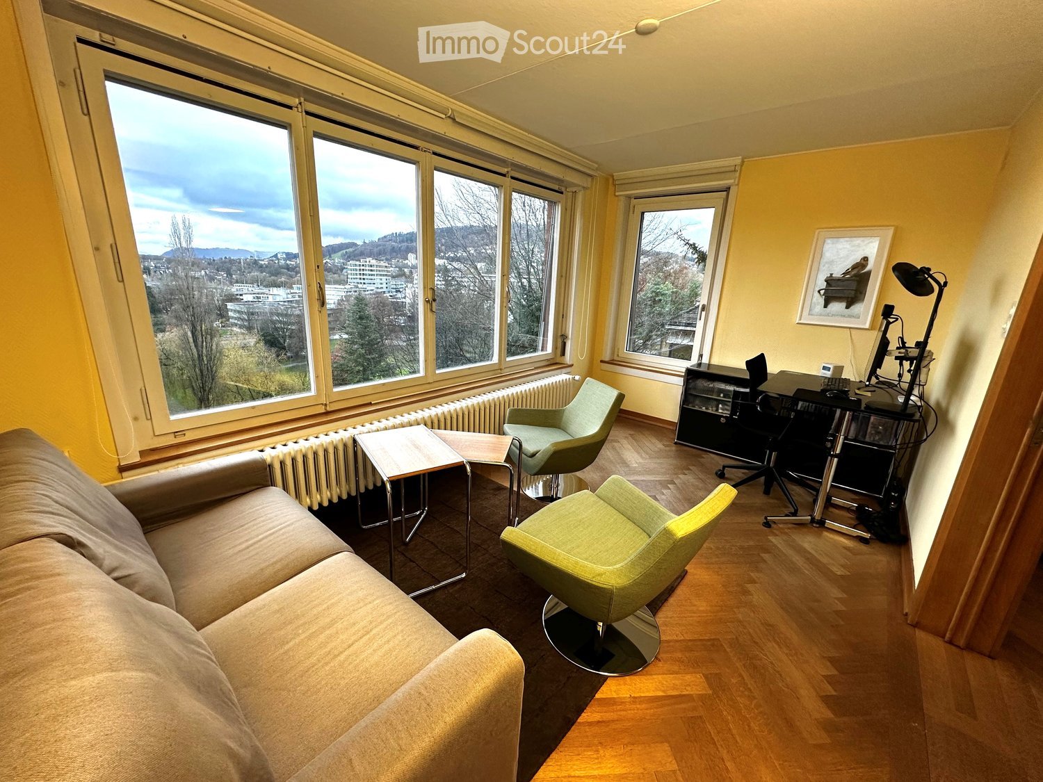 Bright living room with large windows, wooden floor, yellow couch, green chairs, wooden coffee table, wooden office desk, wooden radiator, black picture frame on the wall