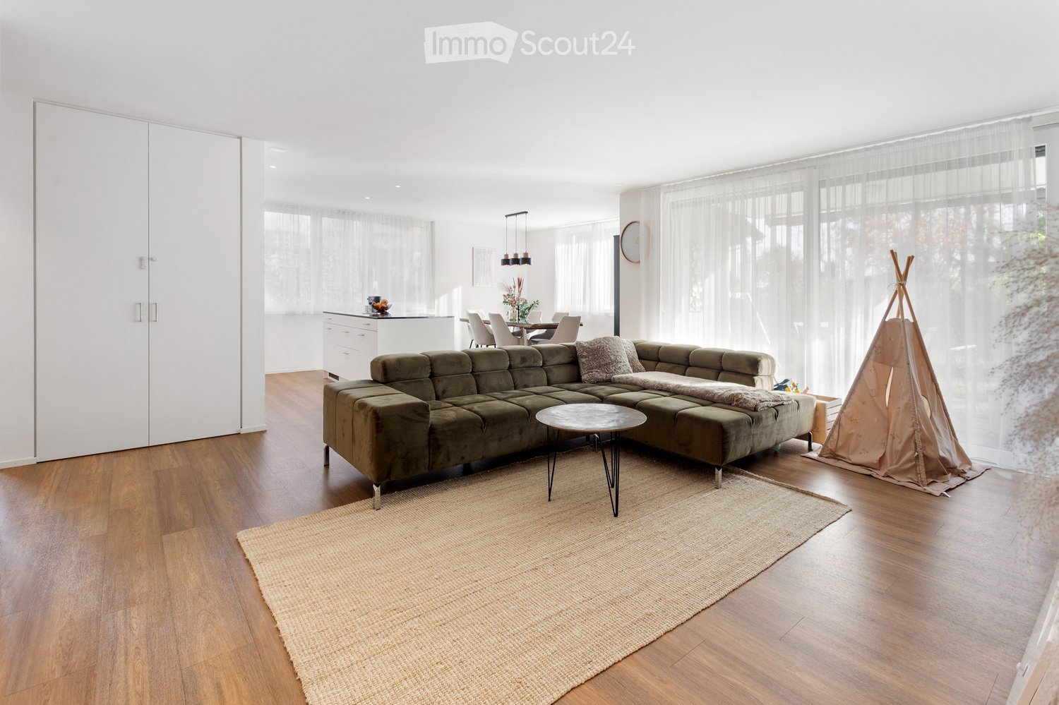 Furnished Room. Modern living room with a green sectional sofa, coffee table, and a small tepee play structure. The room features a kitchen area with a white island and dining table, large windows with sheer curtains, and a wooden floor.