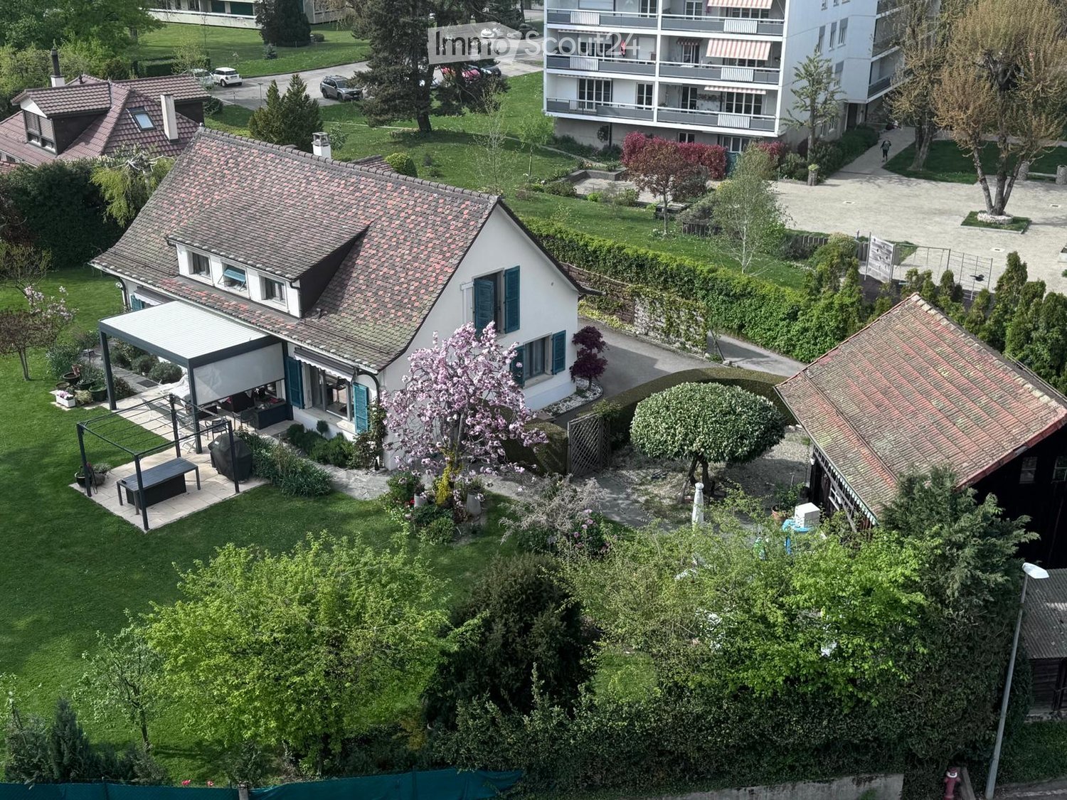 Aerial view of a residential area with a single-family house, garden, patio, and surrounding greenery. The house has a tiled roof, multiple windows, and a covered outdoor seating area. The garden includes lawn, trees, and shrubs.